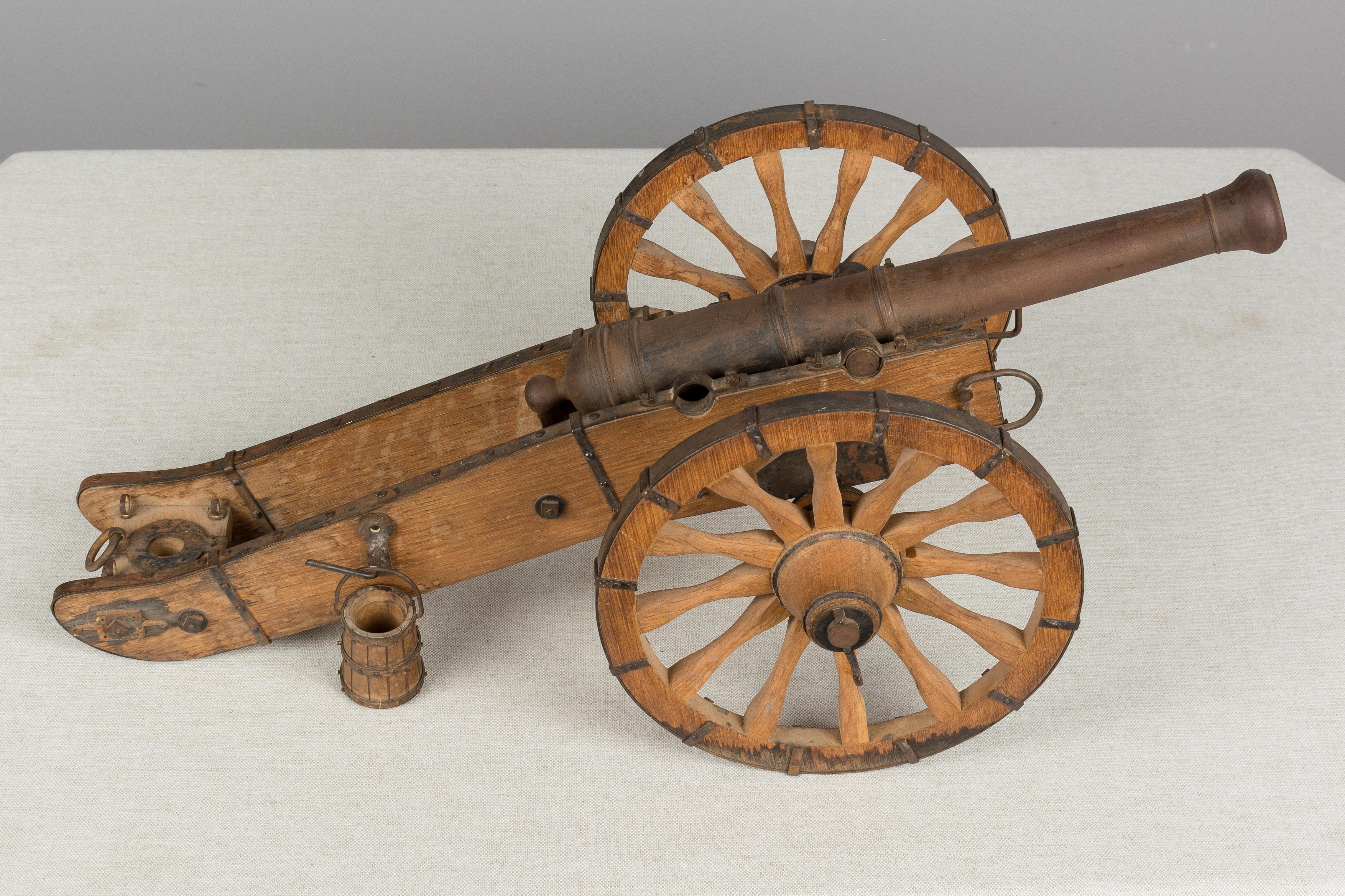 A French miniature model cannon made of iron with wood frame and trimmed in steel with rivets. In good condition with some water damage to wheels and rusty patina to iron. There is a missing bracket underneath that holds one of the wheels. Diameter