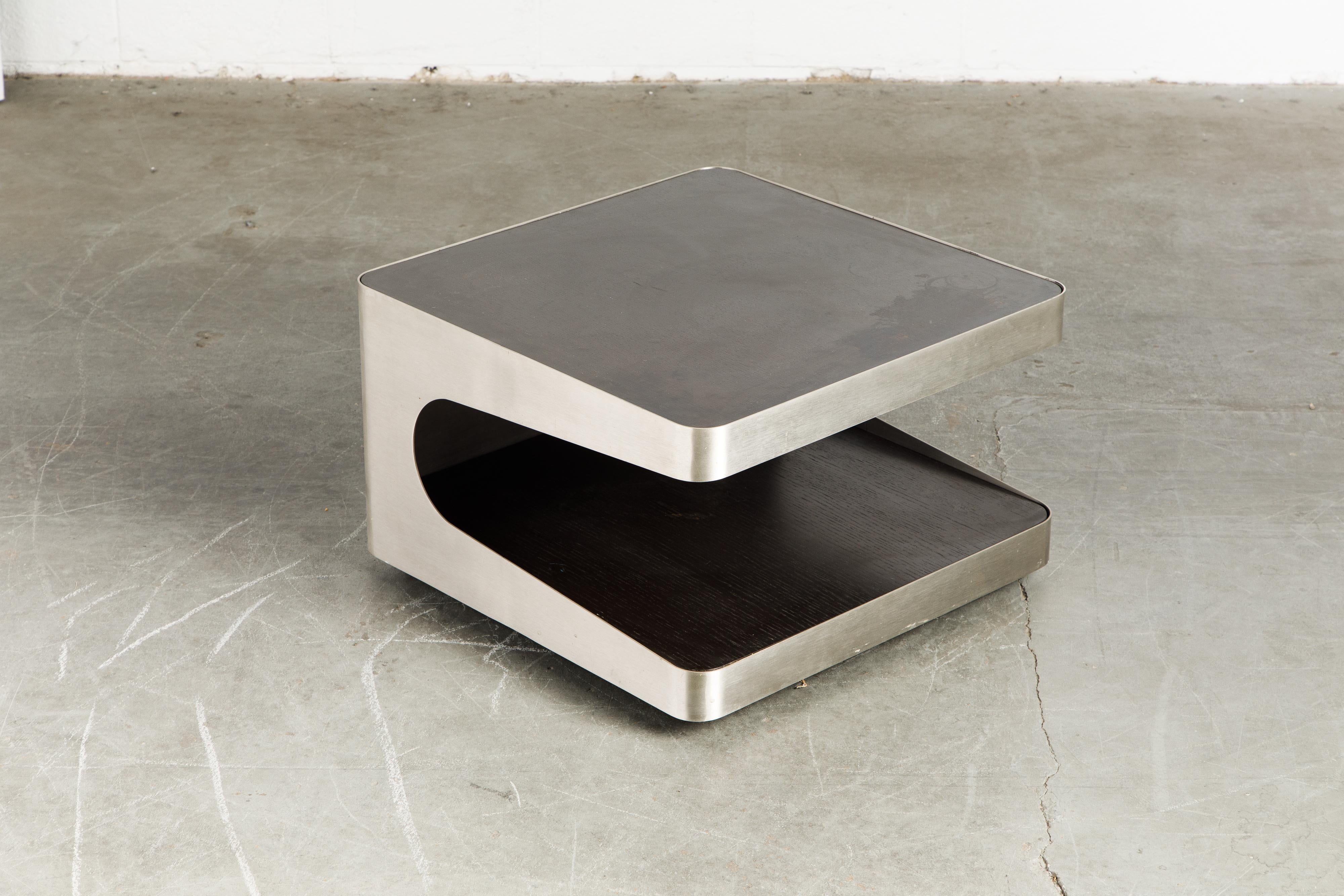 A beautiful French Modern cut steel two-tiered rolling coffee table or end table, circa 1970s. This modernist two-tier table features a brushed stainless steel frame cut into a cantilevered C-shape with two dark brown stained oak wood upper and