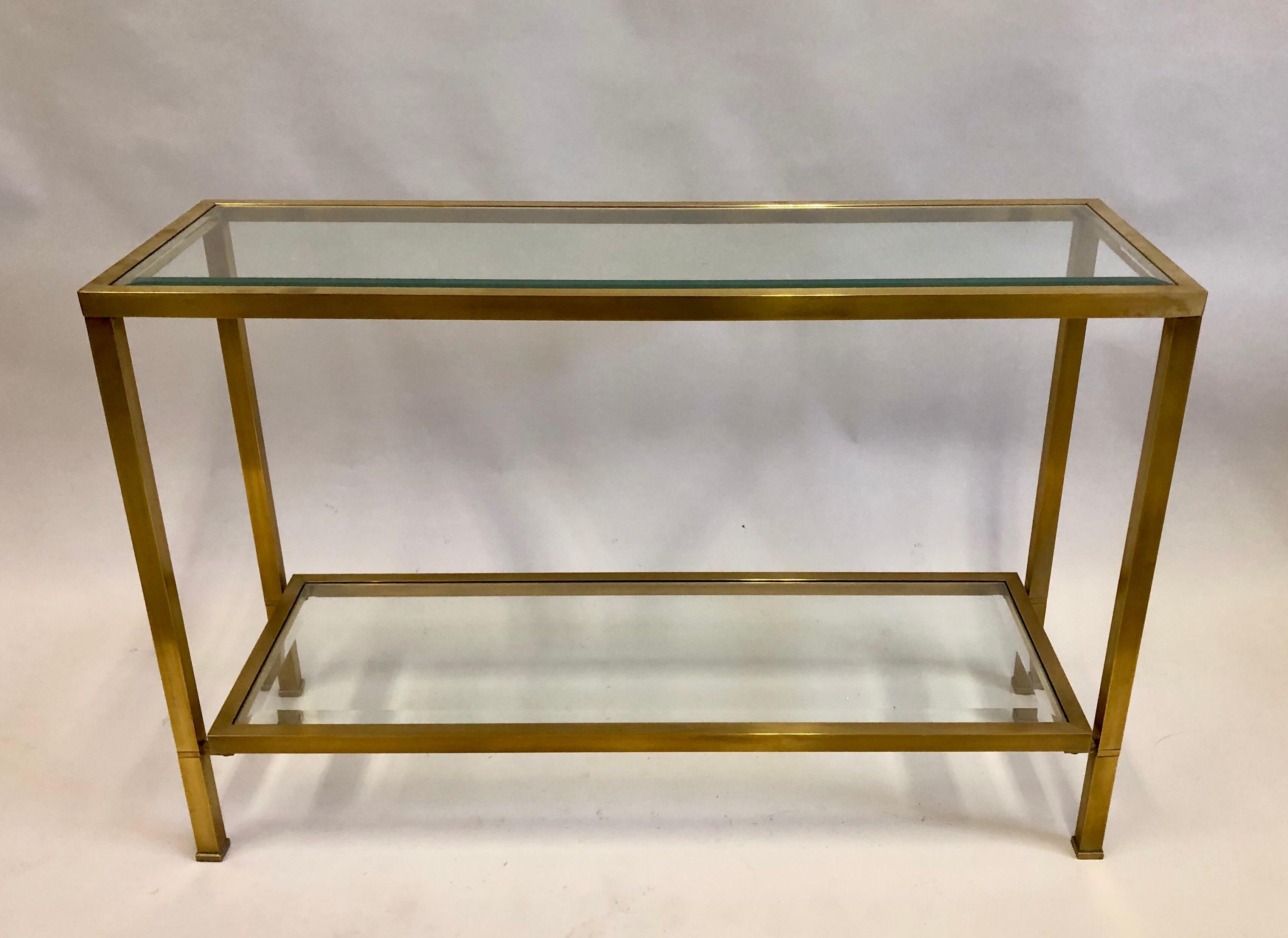 Elegant French Midcentury Solid Brass and glass double level console or sofa table in the Modern Neoclassical Style. The table is attributed to Jacques Quinet and features sober, refined detailing with the exquisite square form sabots that are