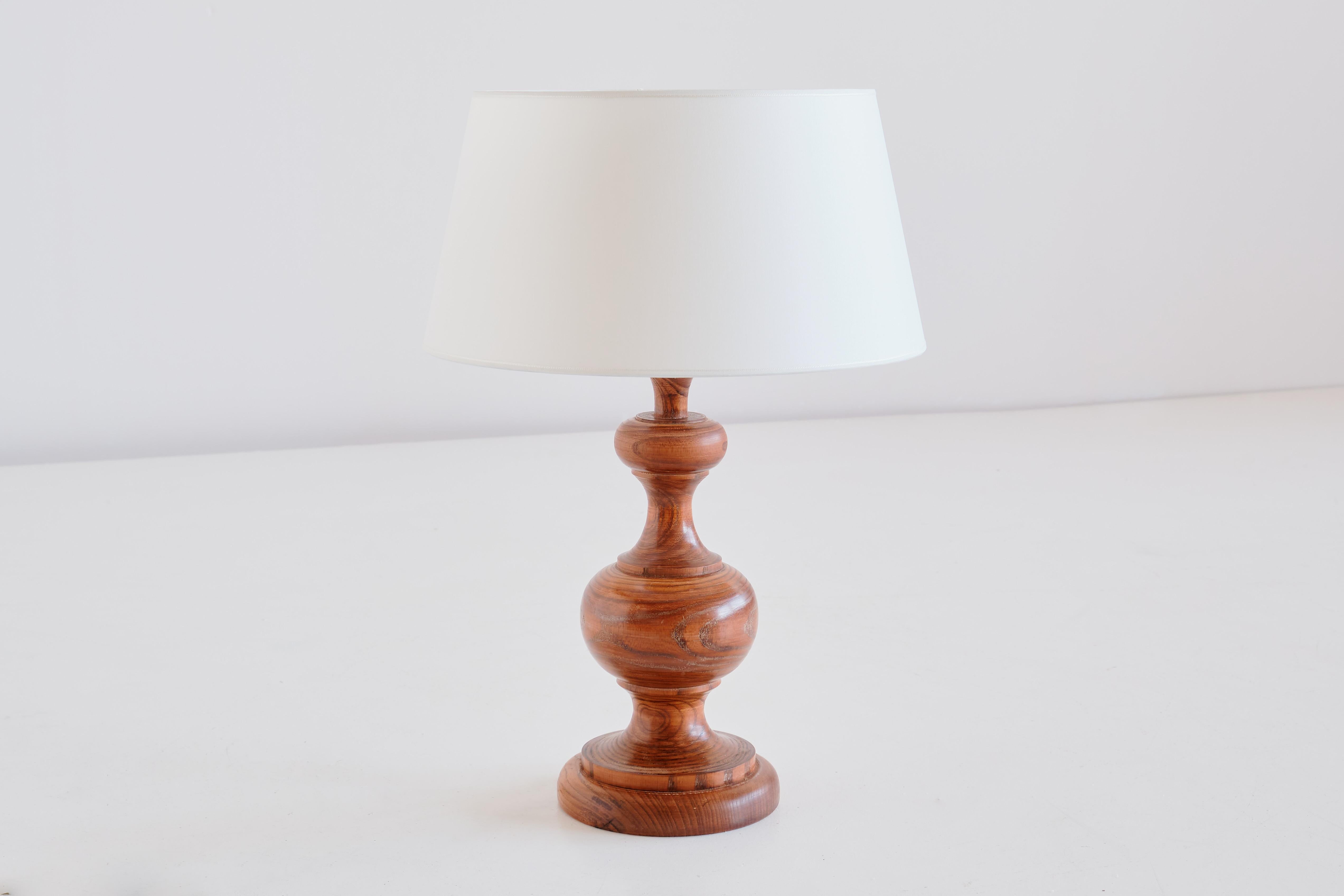 Cette lampe de table rustique et élégante a été produite en France dans les années 1950. La lampe est fabriquée en bois de chêne massif et présente un grain de bois remarquable. Le nouvel abat-jour ivoire en forme de tambour effilé diffuse une