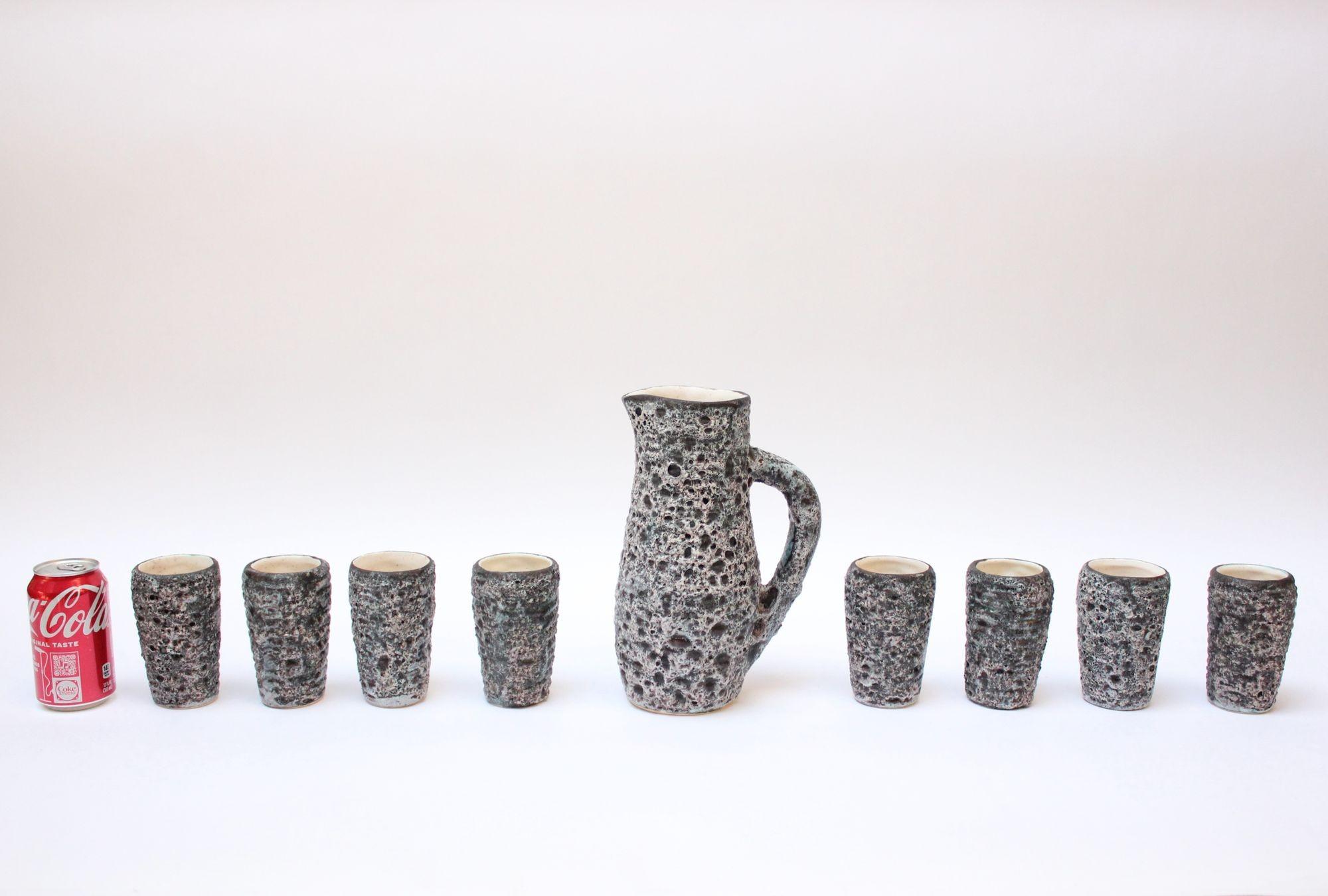 Heavily textured ceramic nine-piece drink serving set including eight cups/tumblers and a single pitcher (ca. 1960s, Annecy, France). 
Fat volcano lava glaze in charcoal and white with subtle hues of turquoise and pink. 
Each piece is signed