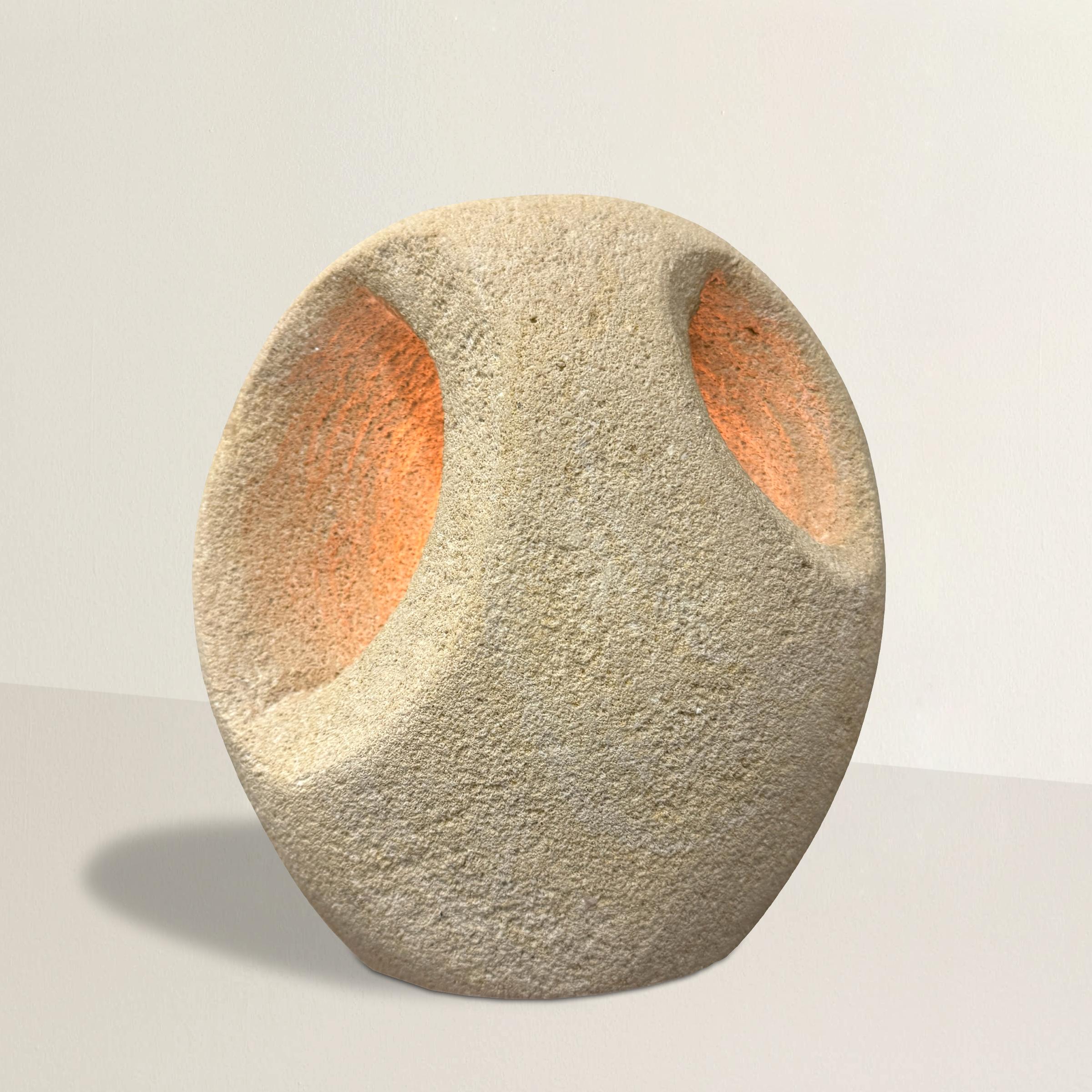 This late 20th-century French Modernist-inspired carved limestone table lamp is a stunning blend of form and function. Its ovoid shape is complemented by two oval voids carved into the front, allowing the soft glow of a single light bulb to emanate