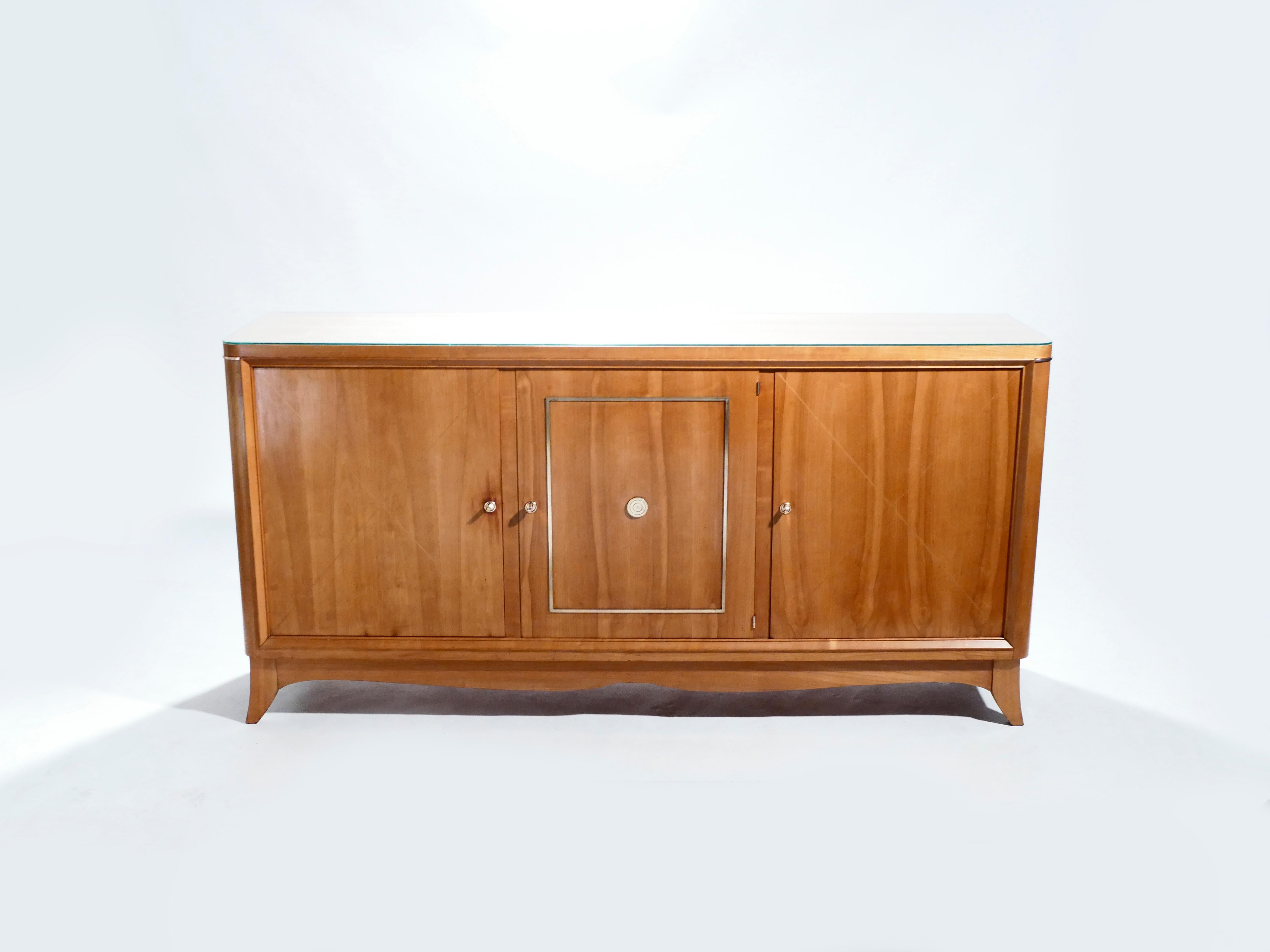 This attractive sideboard features a smooth cherrywood build and bright brass accents. The sideboard is reminiscent of French designer Jean Pascaud or Suzanne Guiguichon acclaimed work. The eye-catching brass decorative knobs on the centre cabinets