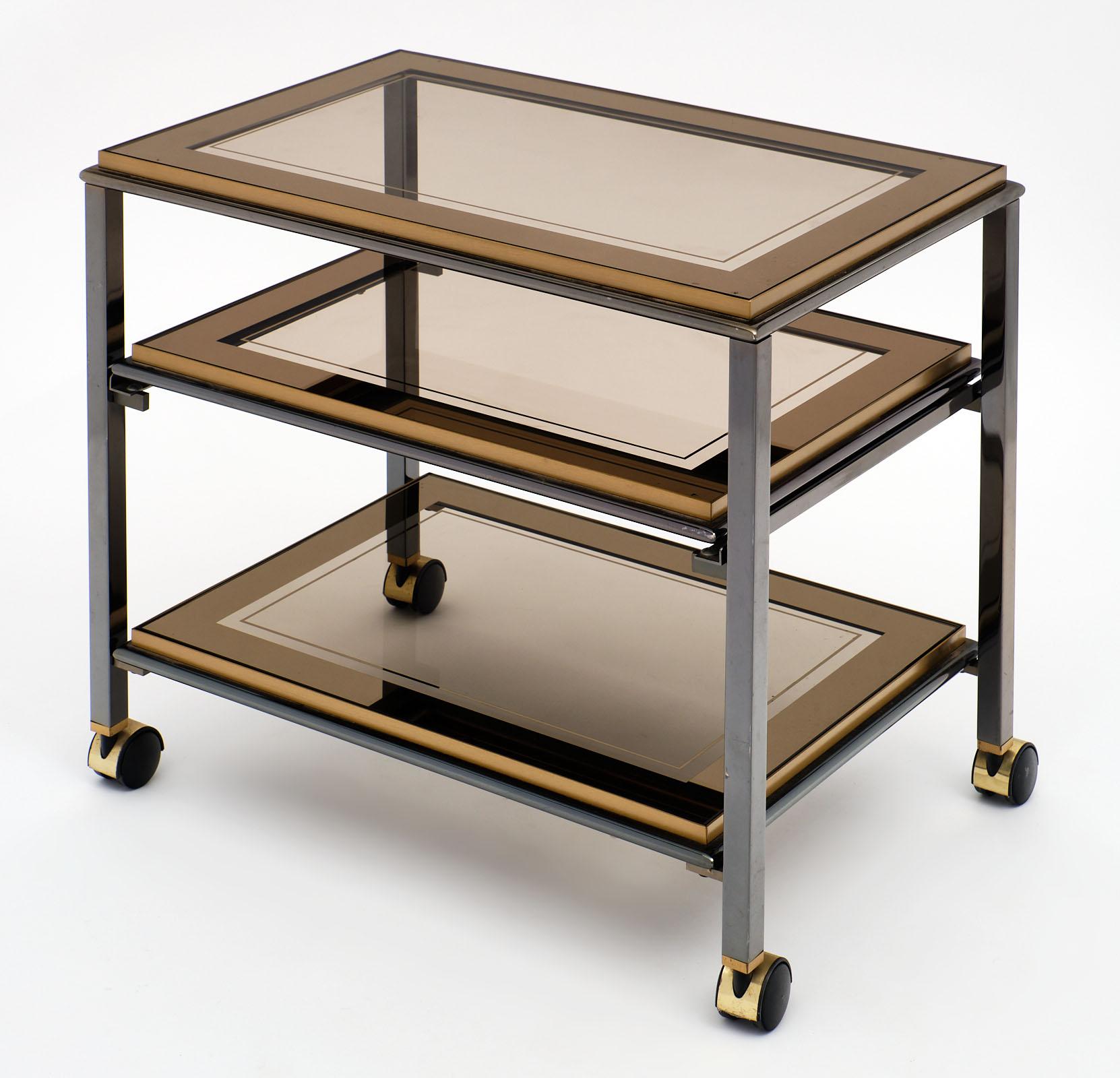 Modernist French side table on casters made of steel and brass. This piece features three shelves and the middle shelf slides forward for functionality. The shelves are smoked glass with the original mirrored banding on the glass.