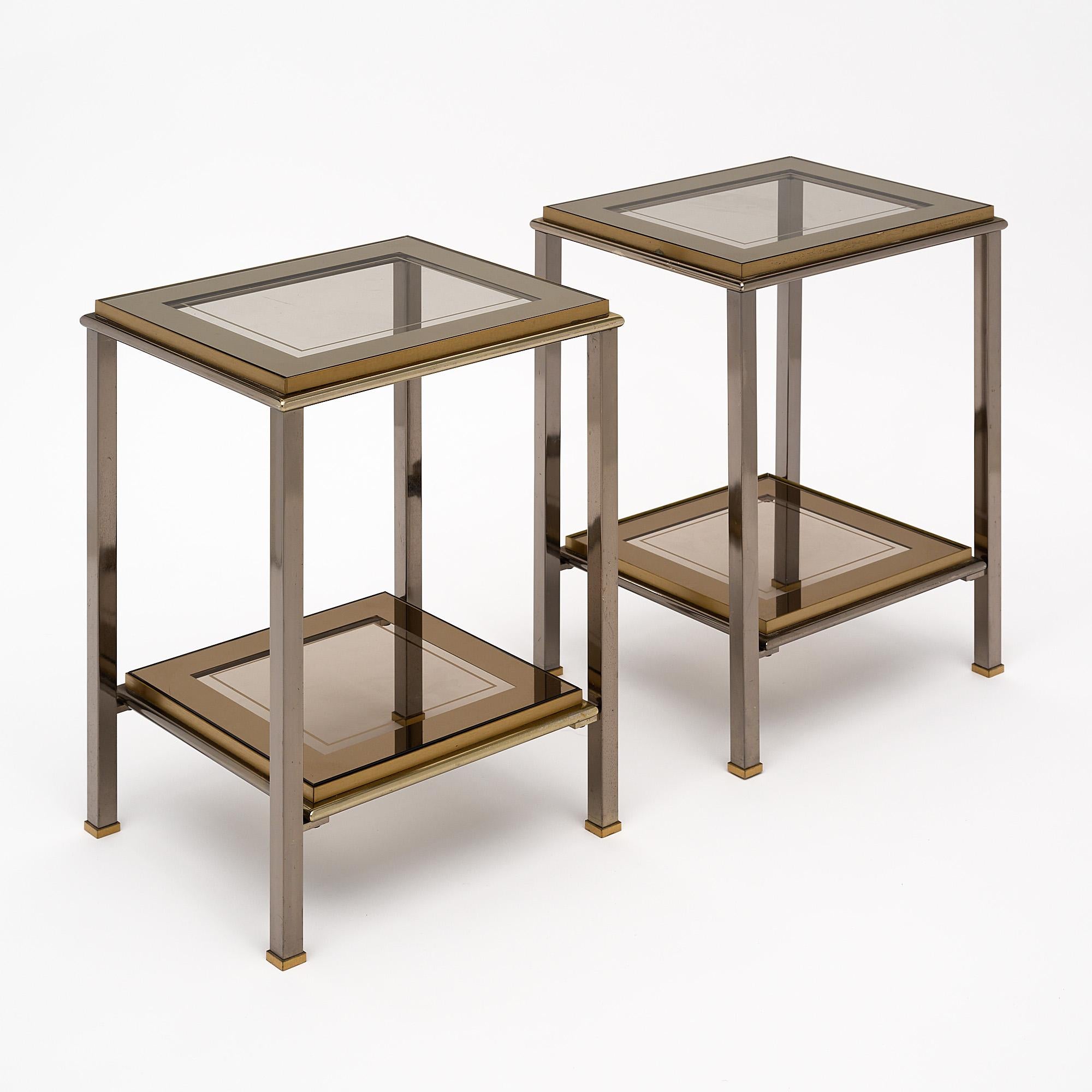 Modernist French side tables made of brass. This pair features two shelves of smoked glass with the original mirrored banding on the glass. The pair has two-toned metal finishes, both brass and a gunmetal toned finish.
