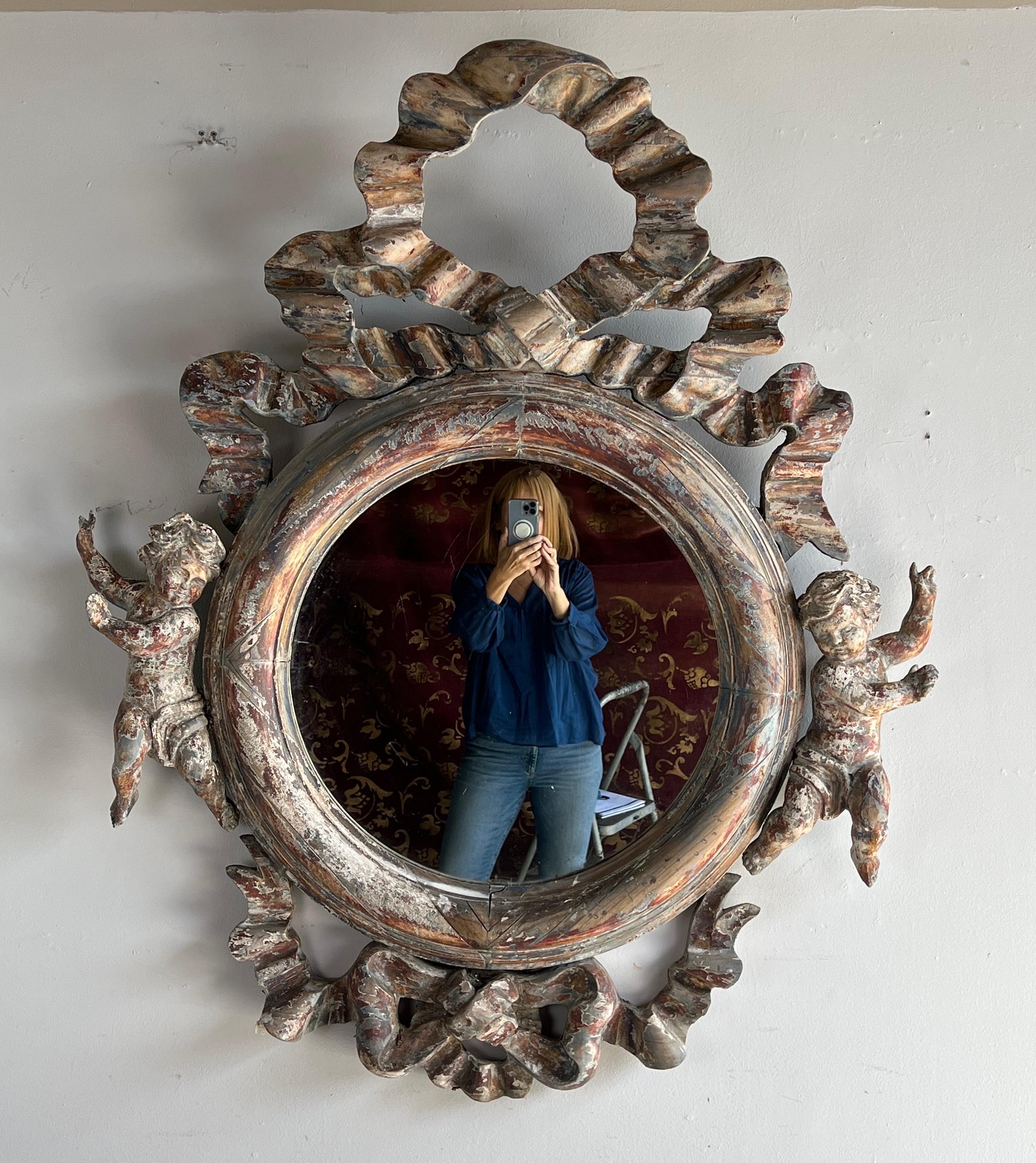 Monumental size French carved wood painted mirror. The mirror depicts a large bow with cascading ribbon and a charming cherubs flanking the sides. The worn painted finish has lovely shades of rust, blue, cream & gray. The natural wood also shows