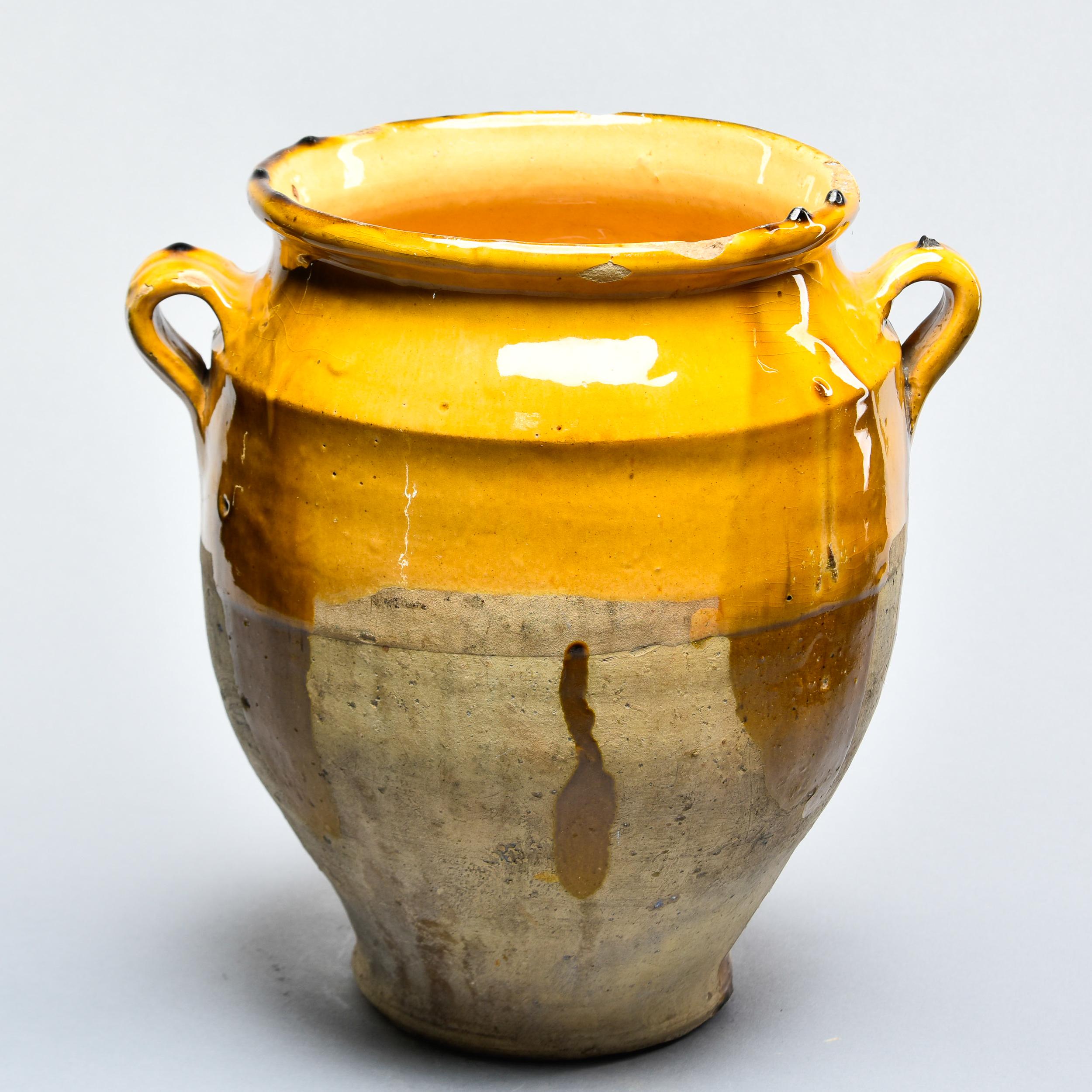 Found in France, this French confit jar dates from approximately 1910. This piece stands 12” high and has the traditional form with a wide vessel body and two handles on the sides with a mustard-colored glaze on the outside top half and fully glazed