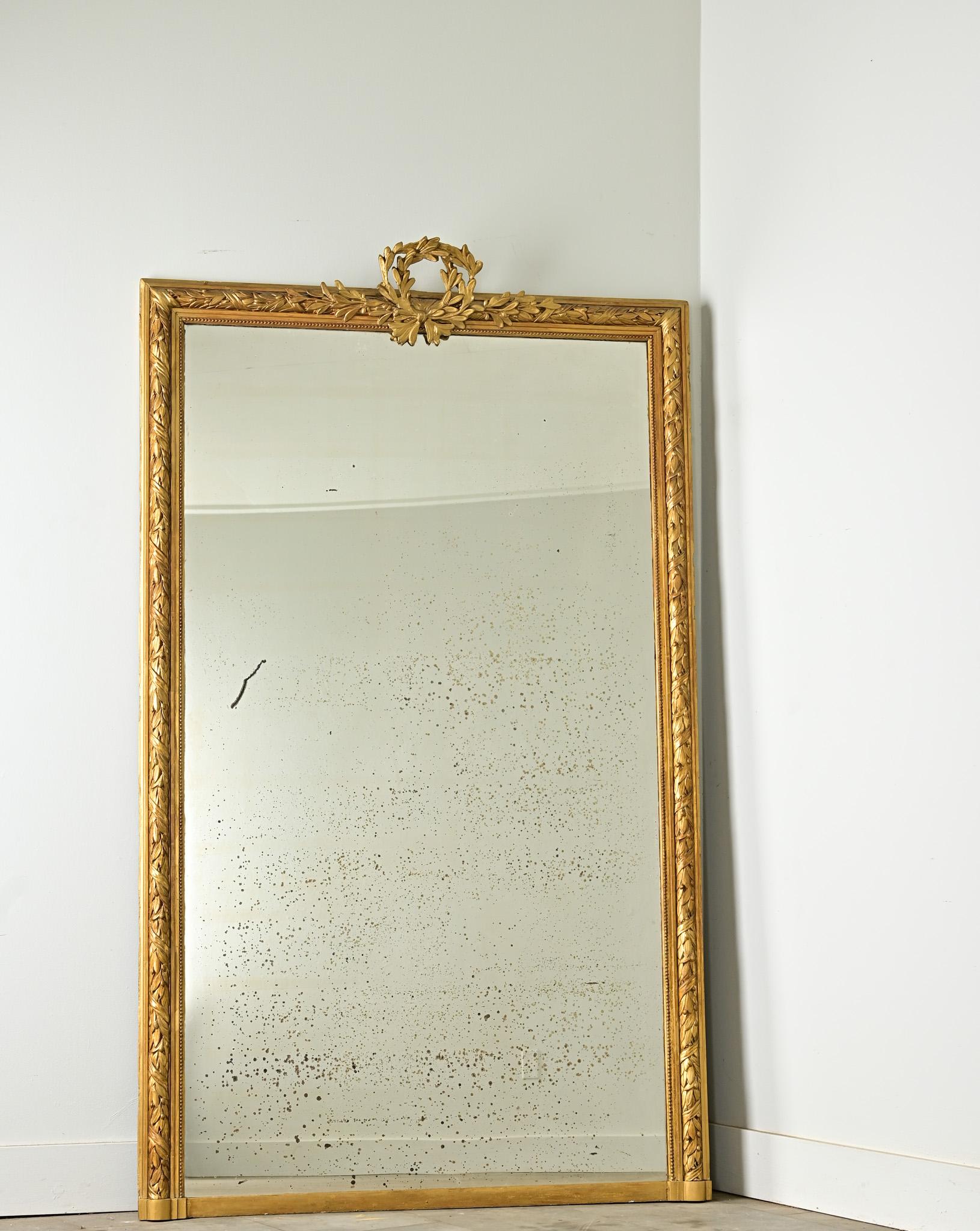 Crowned with an auspicious laurel wreath, this mirror from circa 1870 France will make for an impressive addition to your mantle. Bountiful, leafy vines and twisting ribbon motifs fill the carved giltwood molding, the density of the vegetation