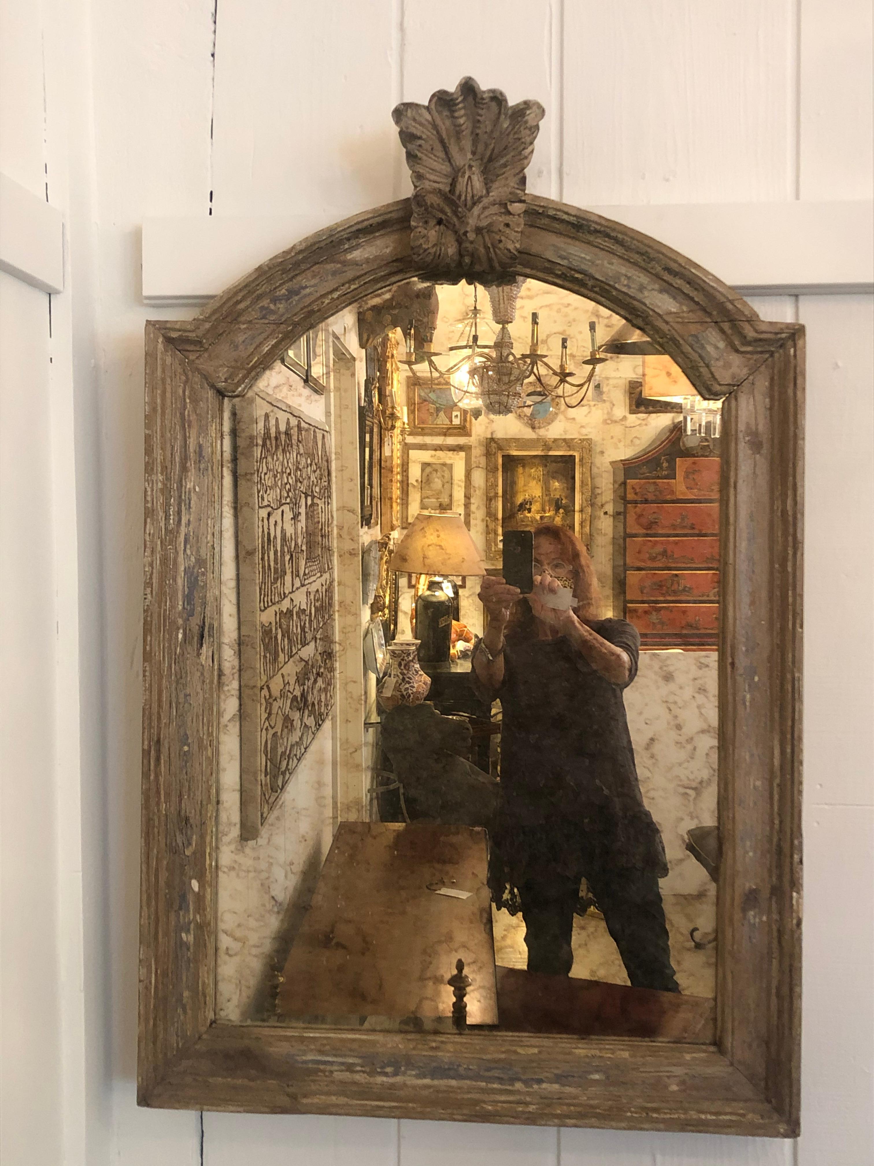 A charming rustic painted and carved wooden mirror having lovely decorative crest at the top and fabulous antiqued mirror. The aged molding is naturally distressed wood with some vestiges of grey, red oxide and cream paint.