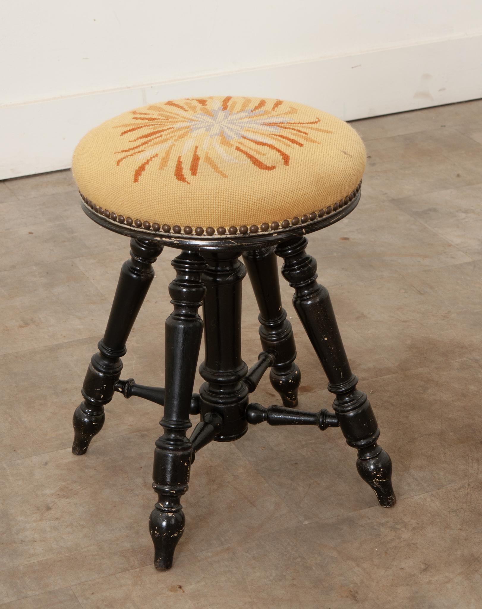 A round French piano stool made of turned wood with an ebonized finish. The top cushion is upholstered with a needlepoint fabric and nailhead trim. The center of the stool has a functioning steel screw to raise and lower the seat height making this
