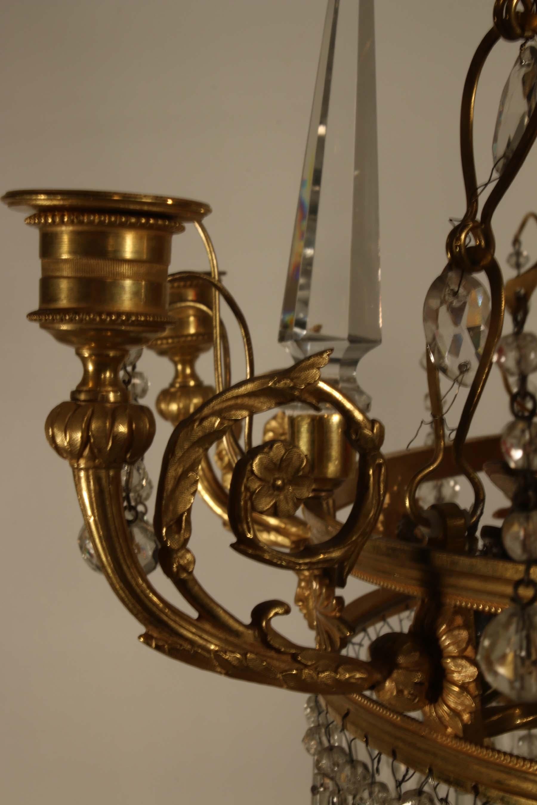 This is a really unusual chandelier, even the chains are set with faceted crystals. They support a gilt bronze circular band crisply pierced with masks and acanthus and with six candle holders and six crystal spires. Suspended below are a mass of