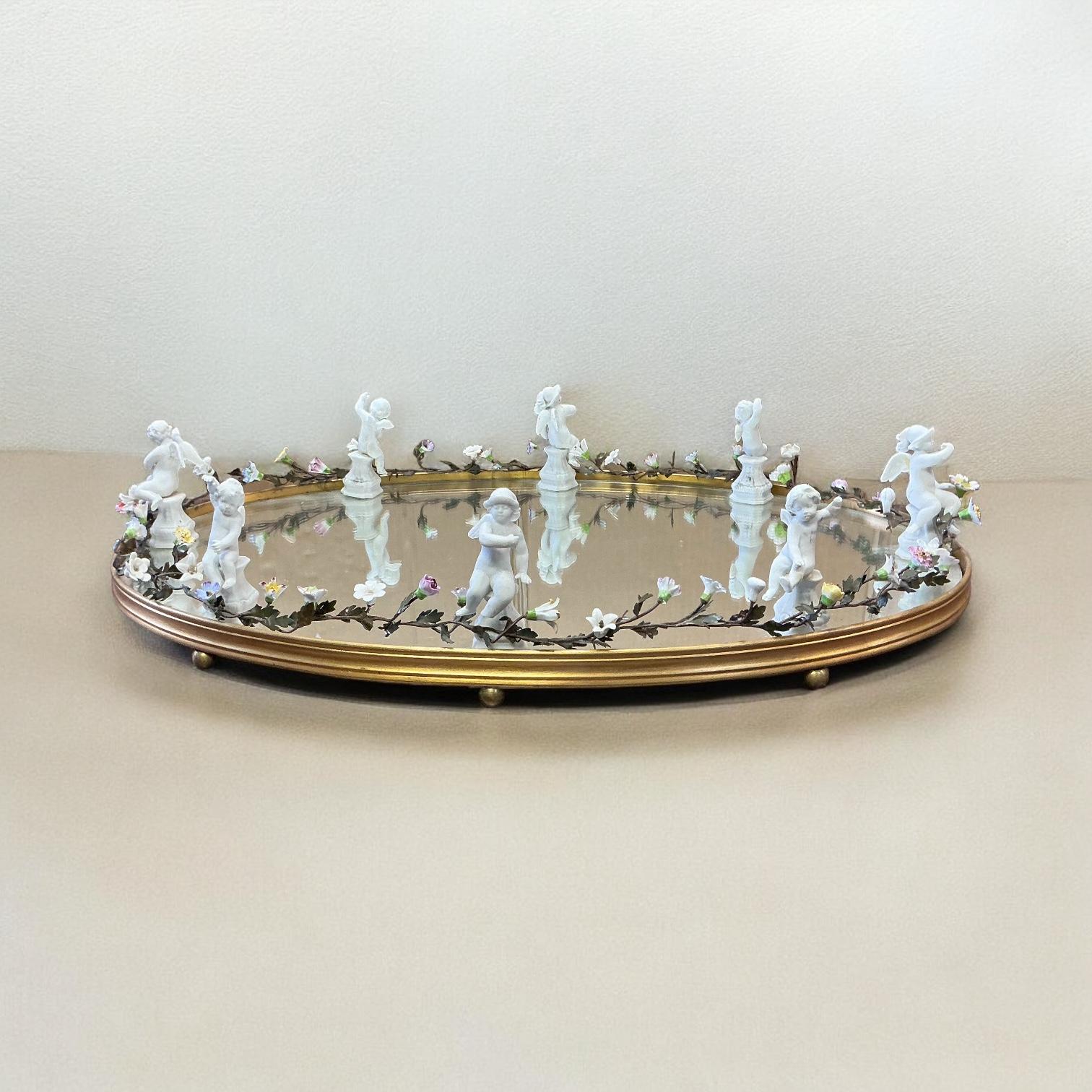 This is an early French neo-classical style mirrored plateau or tray. The mirror is resting on a gilt bronze base. The bisque cherubs or puttis frame the tray along with a garland of porcelain flowers. It is unmarked.
