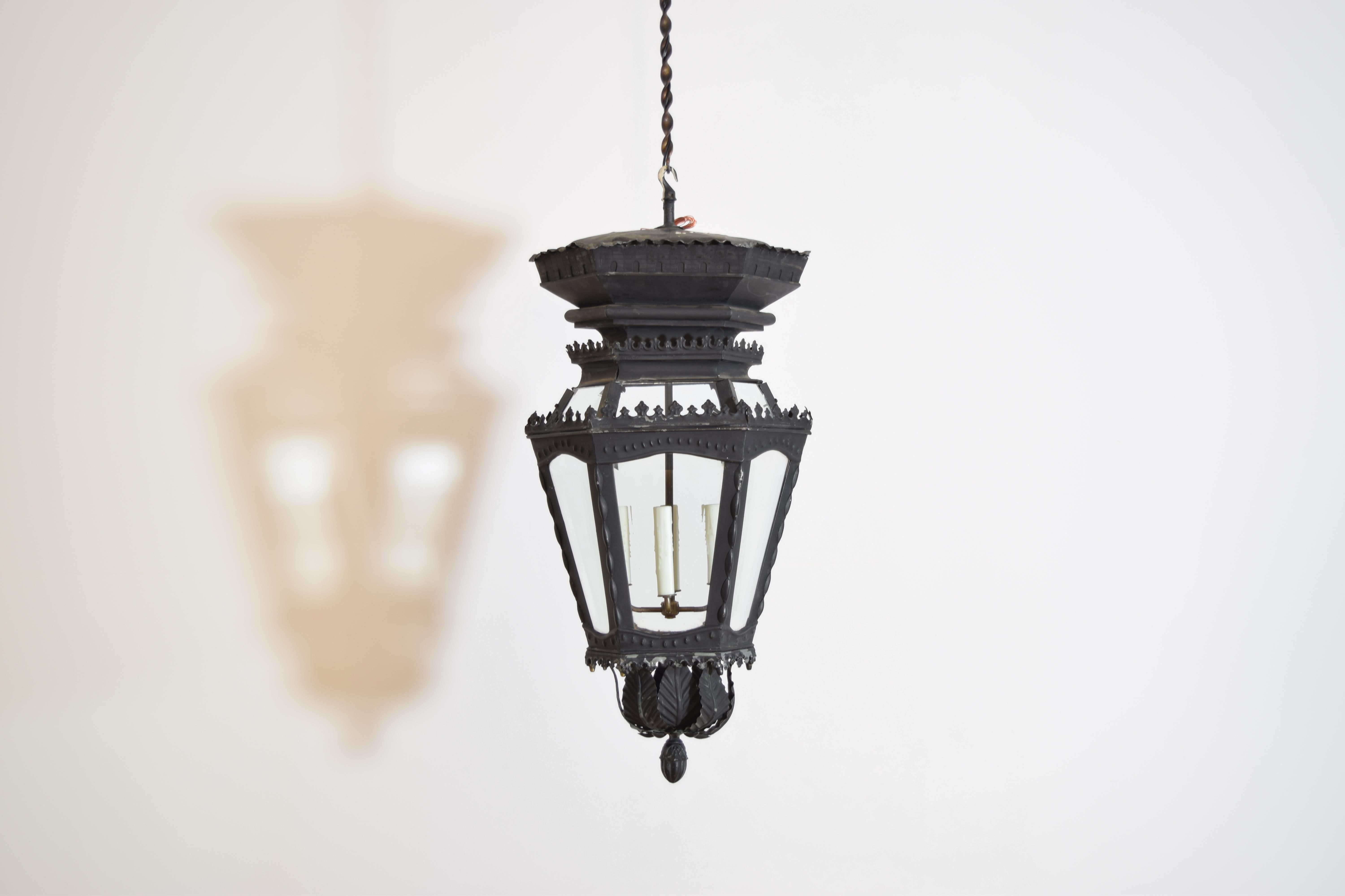 this unique lantern, early 20th century but possibly earlier, retains its original beveled glass panels, the top with impressed crenellated detail, the tapering hexagonal body with separate side and top glass panels, the bottom with a tole leaf
