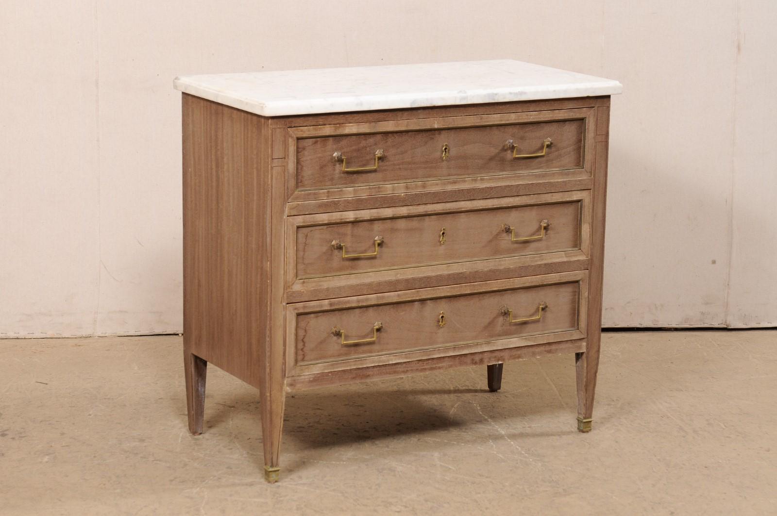 A French neoclassical-style three-drawer commode with brass accents and white marble top. This vintage chest from France features a rectangular-shaped marble top which slightly overhangs the case below which houses three graduated drawers, a