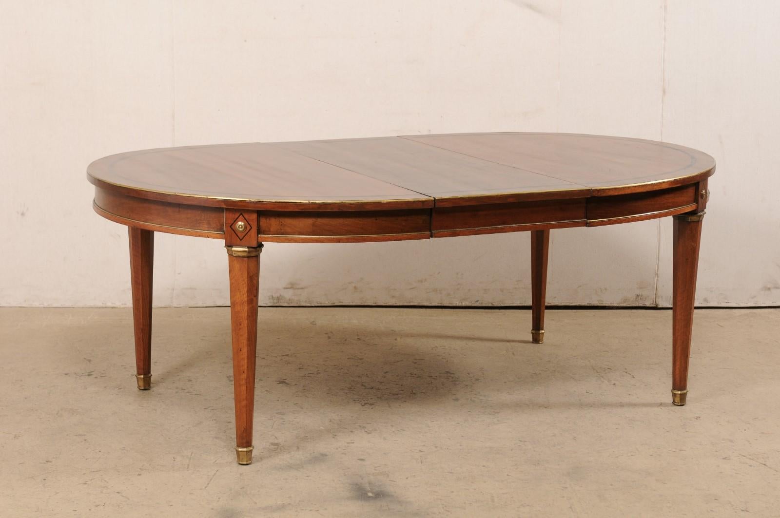 A French neoclassical style table dining table. This vintage table from France has an oval-shape with an oval inlay adorning its top. Brass trim outlines the top edge as well as the clean lines of the skirt about the perimeter. Diamond shaped inlays