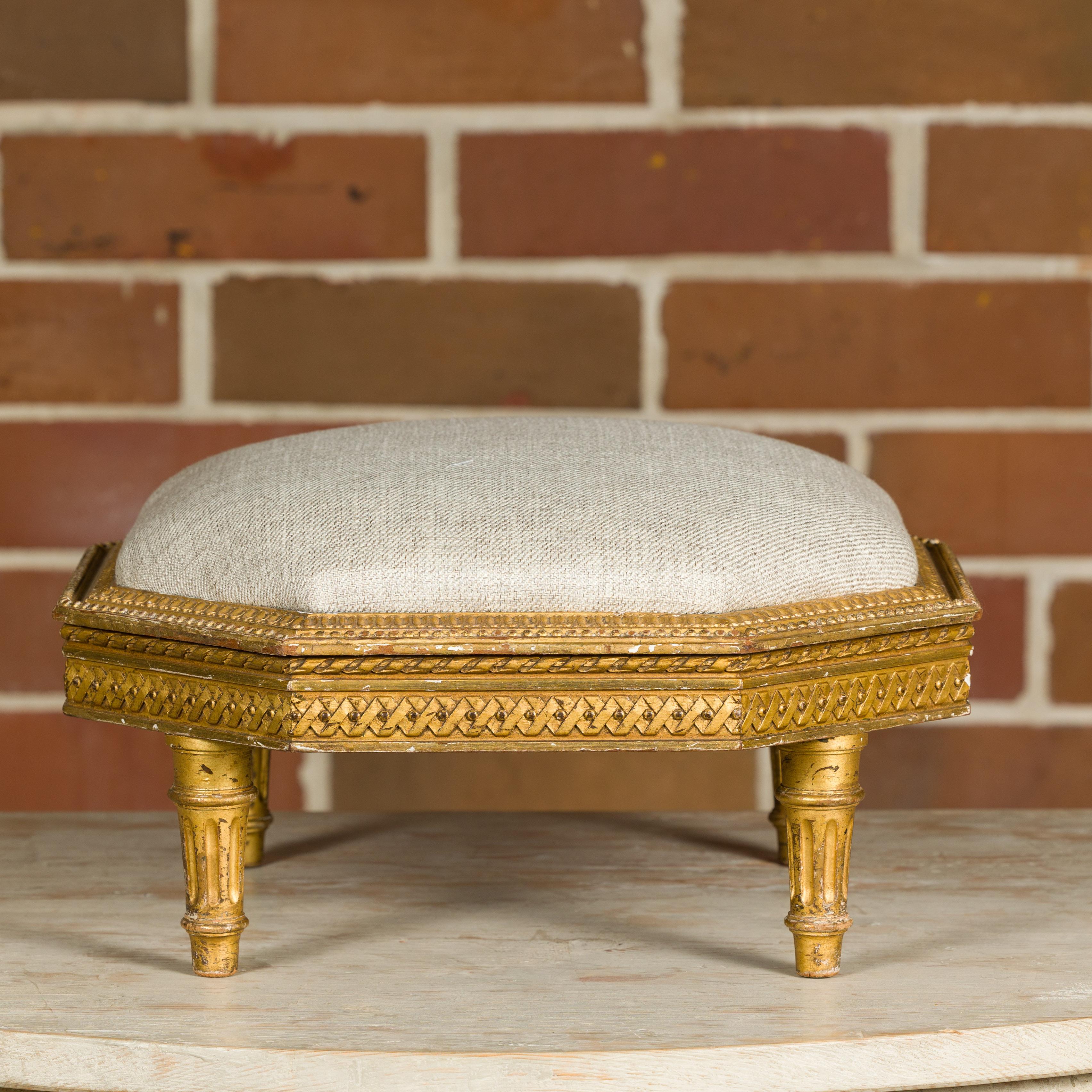 A French Neoclassical carved giltwood footstool from the 19th century with custom linen upholstery. This exquisite 19th-century French giltwood footstool is a testament to timeless craftsmanship and refined elegance. Its octagonal frame is adorned