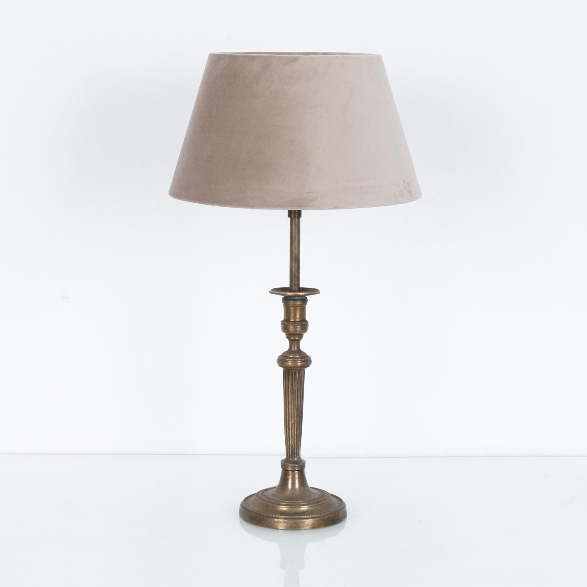 A repoussé brass lamp from France, circa 1900. A fluted, embellished column gives a neoclassical feel. The lampshade, a soft beige, harmonizing with the muted tone of the metal. Stately yet understated, this table lamp provides an elegant accent,