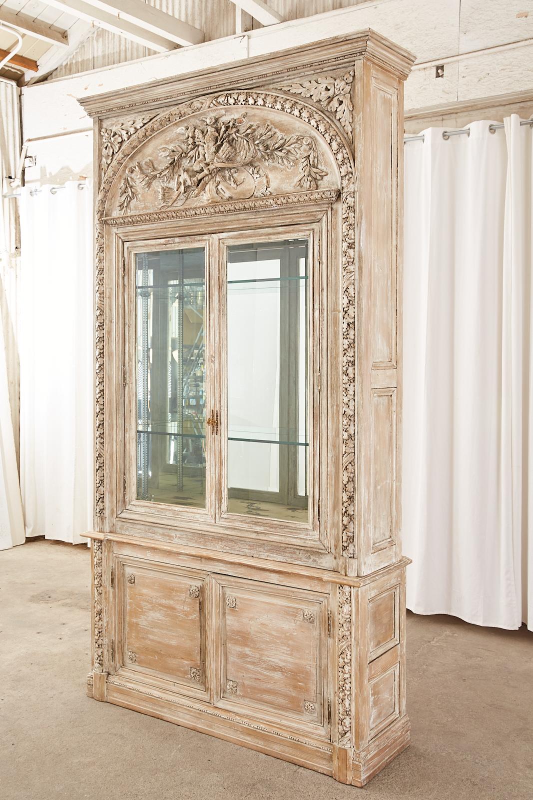 Magnificent French bookcase or display cabinet crafted in the neoclassical Louis XVI taste. The two-part case features extensive relief carved decoration with an oak foliate and acorn motif. Constructed from pine with a cerused or white washed