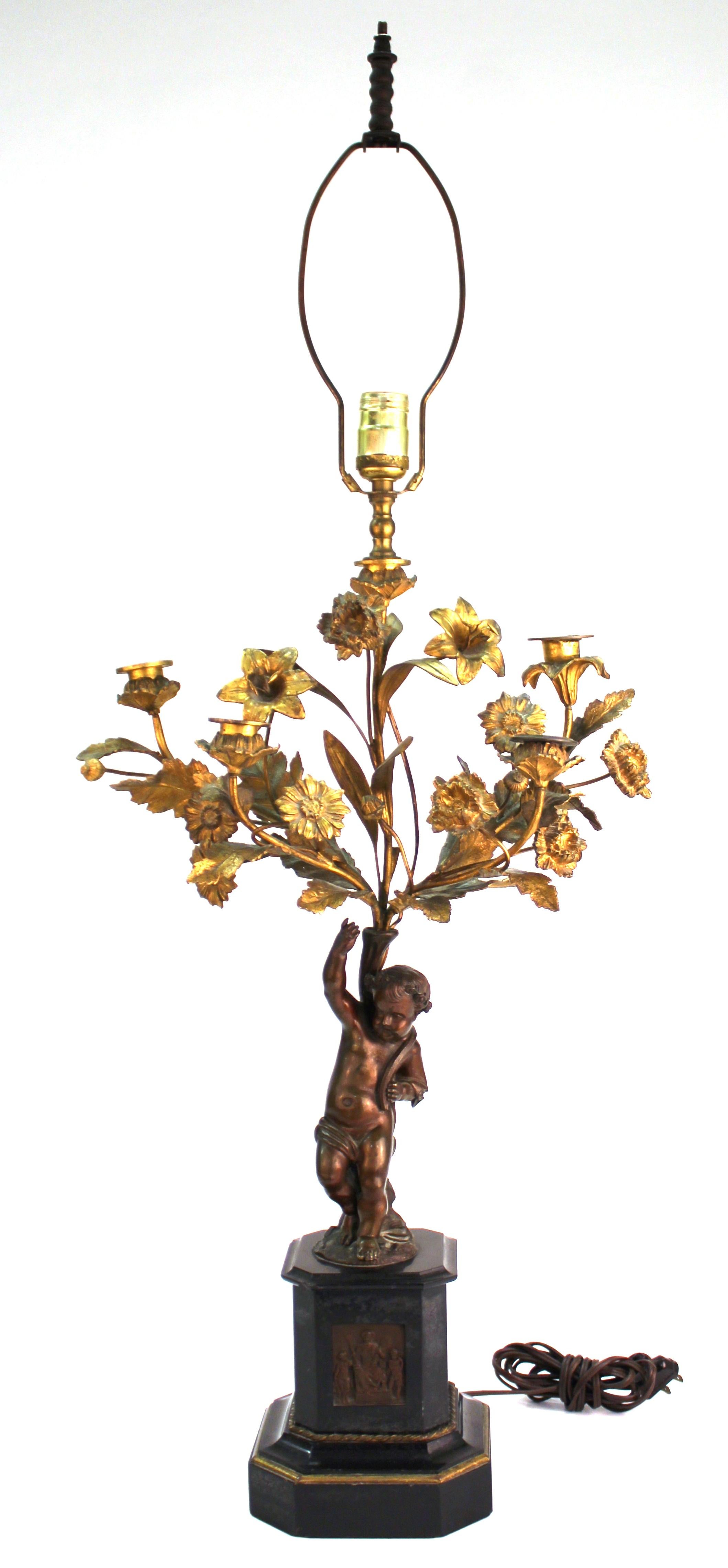 Pair of French neoclassical Revival table lamps with bronze cherub leaning against a floral bush in gilt bronze, atop a marble pedestal base with inserted bas-relief plaque of a seated woman with children flanking her. The pair is in great vintage