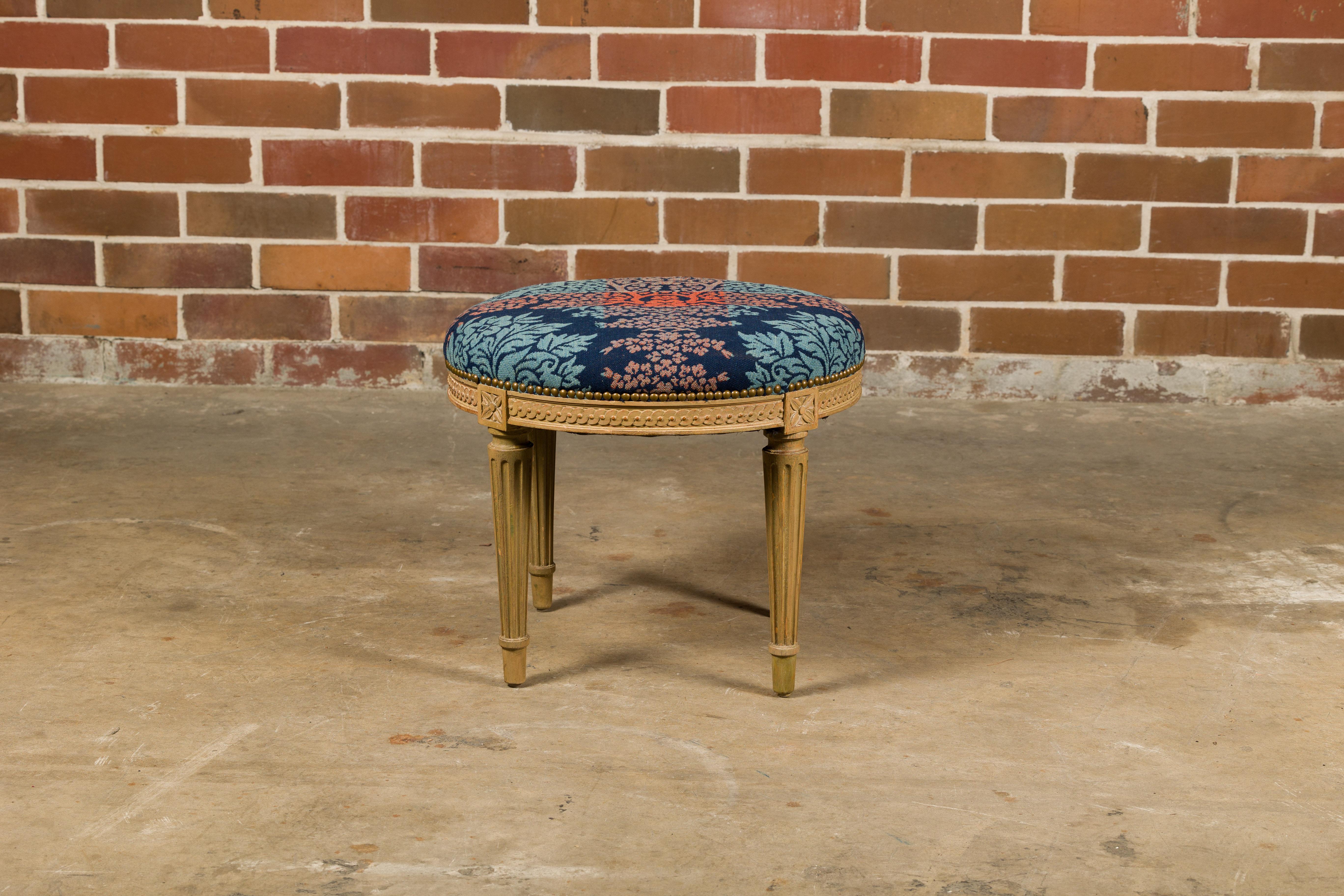 A French Neoclassical style painted stool from circa 1920 with circular upholstered seat, guilloche carved apron and fluted legs. This French 1920s stool, with its circular upholstered seat, guilloche carved apron, and fluted legs, beautifully