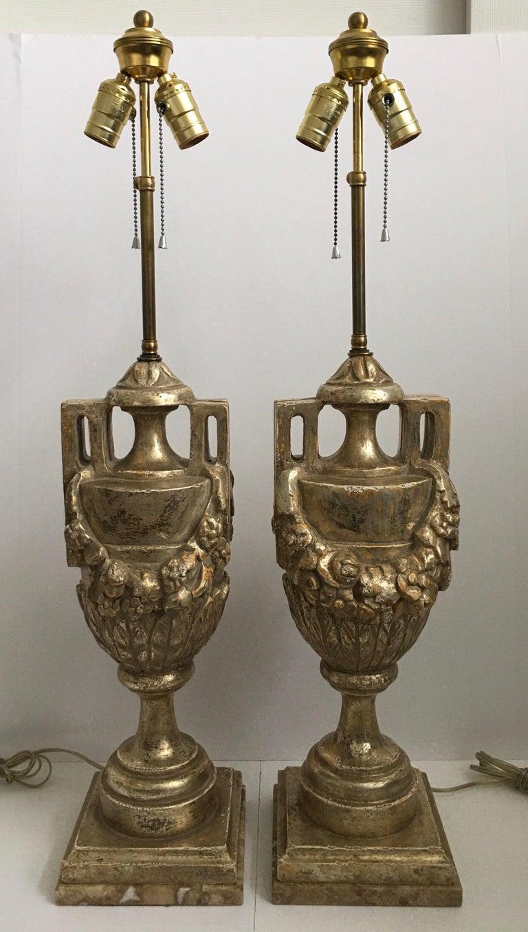 Highly detailed pair of French neoclassical style silver leaf giltwood urn lamps featuring hand carved draped foliate garland motifs. These Louis XVI style lamps are mounted on marble plinth bases and have adjustable riser finials. Each lamp is