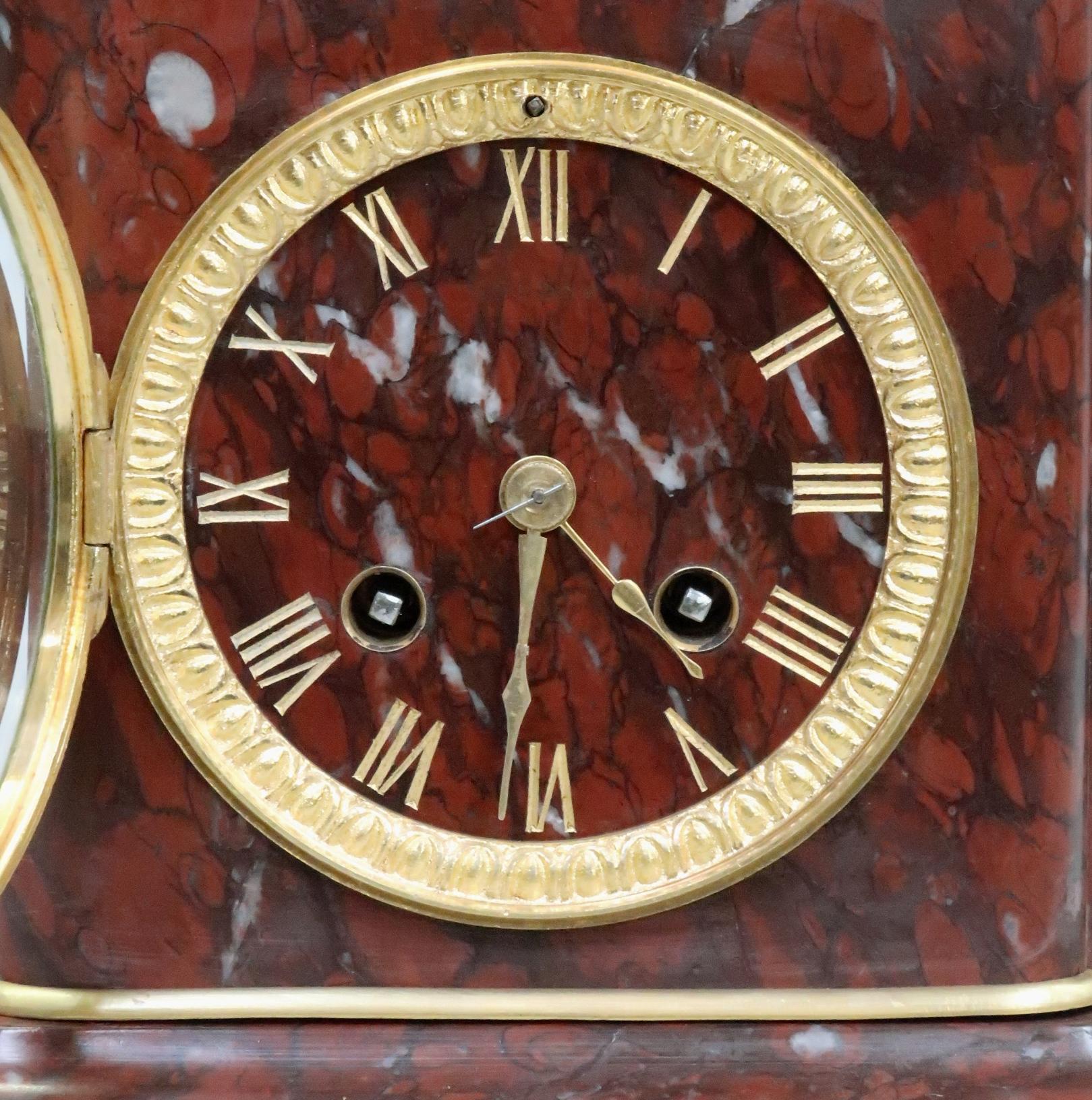 An extremely good quality French neoclassical style deep rouge griotte marble mantel clock with urn to the top and bronze gilt masked mounts stood on paw feet. The clock has a decorative brass bezel with etched gilt Roman numerals and a French eight
