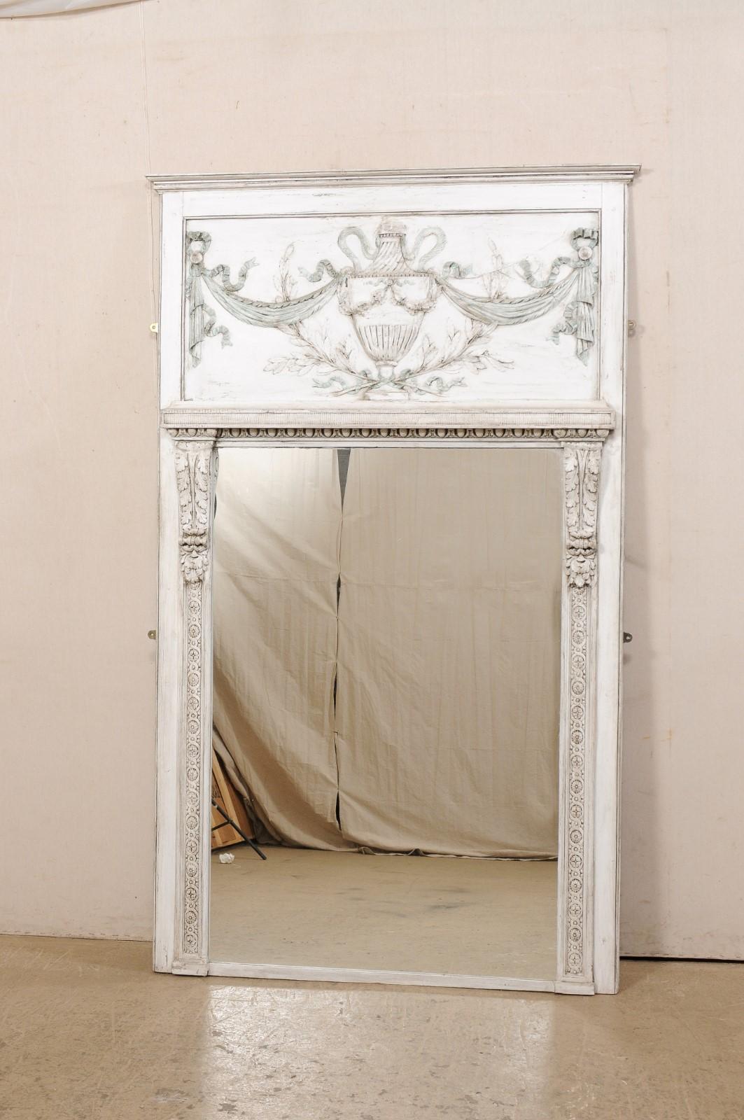 A French Neoclassic-style painted wood trumeau mirror. This vintage mirror from France features the carved-wood plaque top, reminiscent of trumeau design, with carved urn presented prominently and adorn with draped swags, cascading ribbons, and