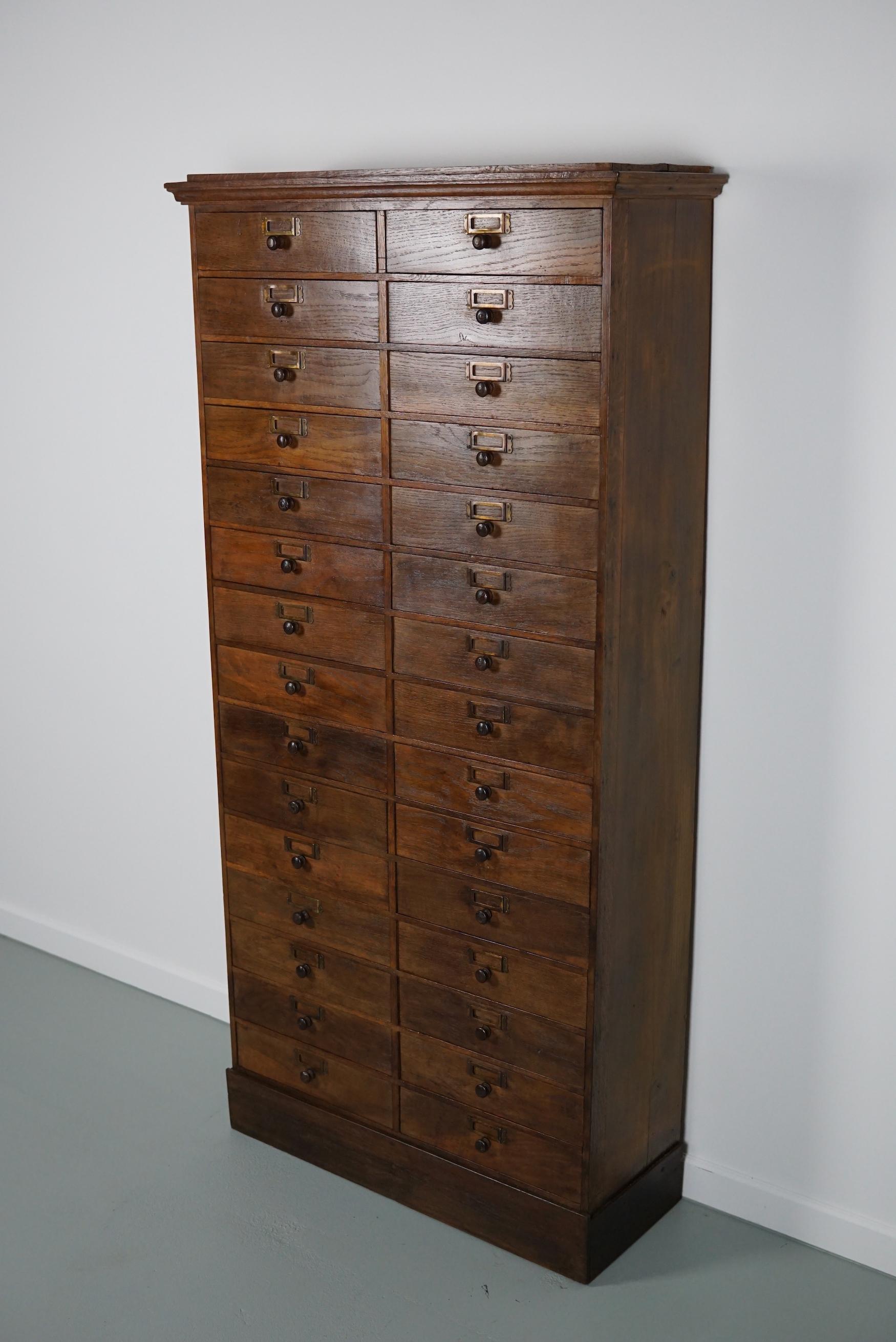 This cabinet was made in France from oak and pine in the early 20th century. It features 30 drawers with oak knobs and brass card holders. It was used to store parts in a watchmaker / jewelry workshop.
