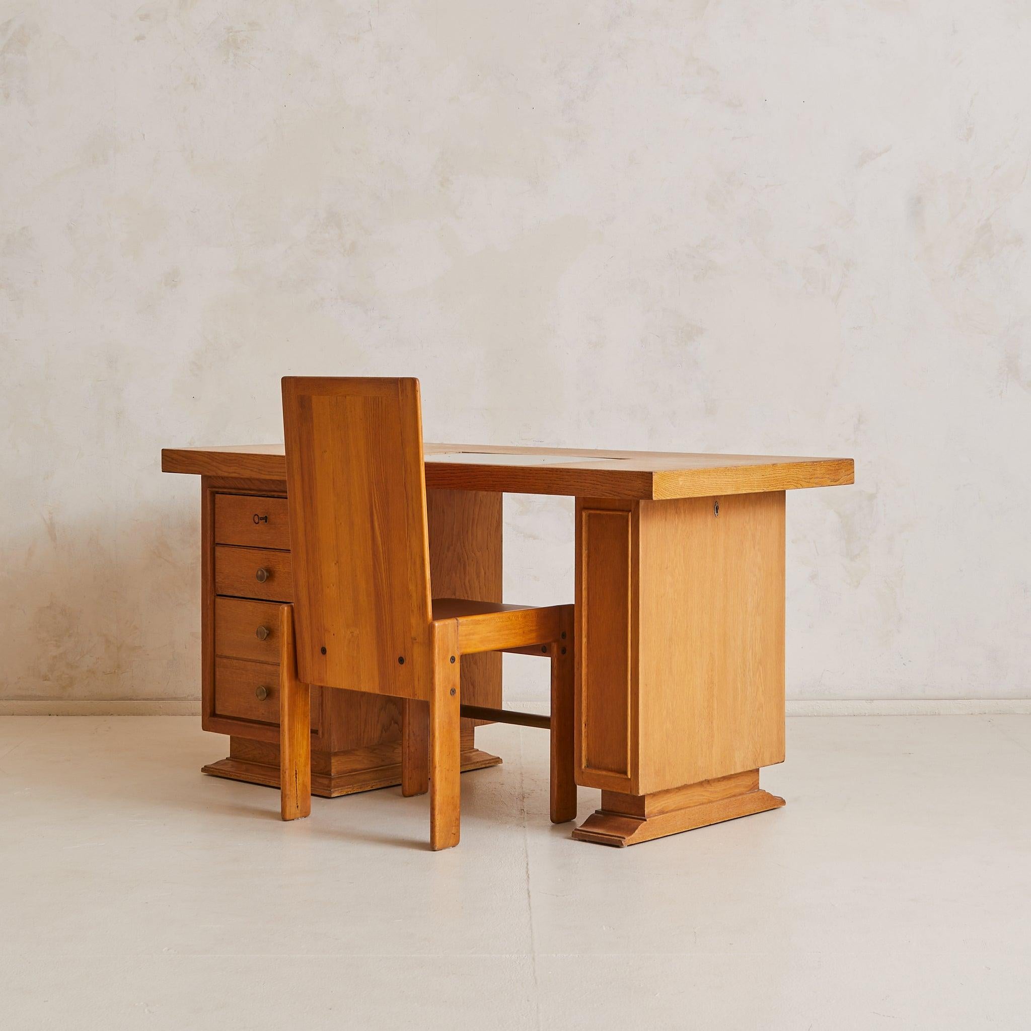 A 1950s French architect’s desk, crafted in solid oak with a warm tone and expressive grain. The rectangular top floats above two pedestal bases on stepped plinths, joined by a single brass stretcher. On one side, a bank of four drawers—complete