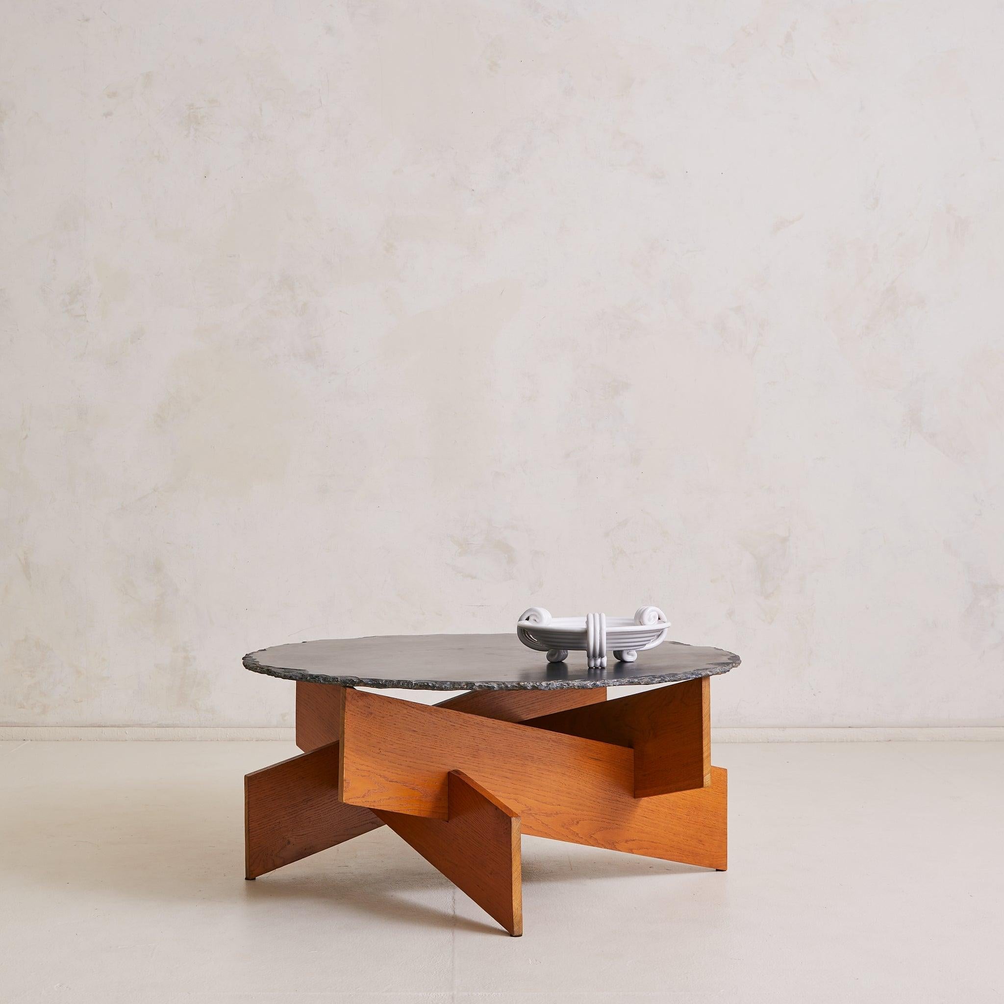 Trouvée dans le sud de la France, cette table basse des années 1970 est une composition de poids et de géométrie. Un plateau rond en ardoise, doucement patiné avec un bord brut, repose sur un socle en chêne emboîté, dont les plans sont inclinés