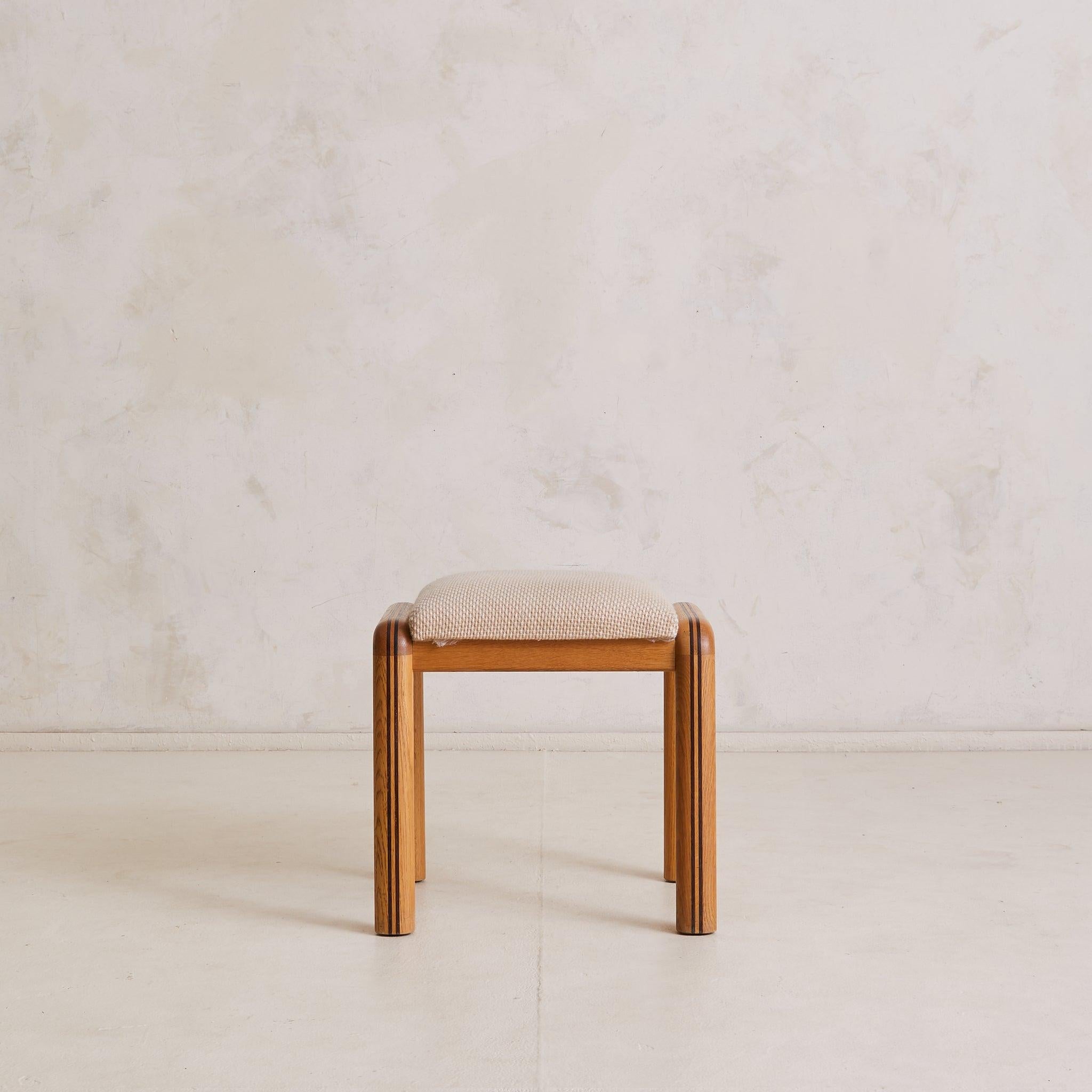 Handmade in France in the 1960s, this oak vanity stool is defined by soft corners and subtle detail. Rounded legs curve fluidly into the frame, accented by deeper-toned wood inlays that complement the warmth of the surrounding oak. The seat, lightly