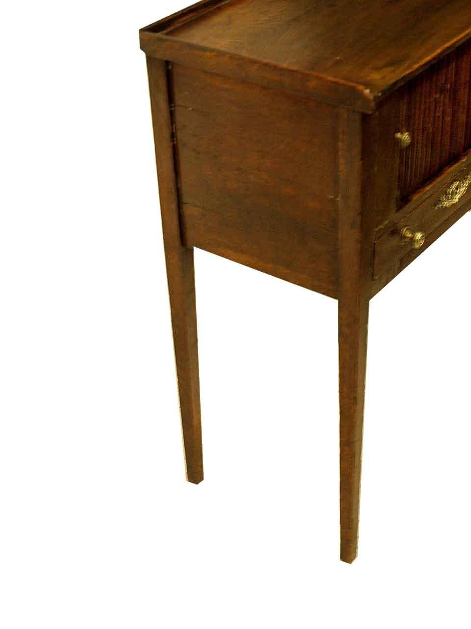 French oak tambour stand, the top has a .38'' gallery around the edge; the front features a sliding tambour door revealing open interior, single drawer with brass knobs and escutcheon; the slender legs are nicely tapered; original finish.