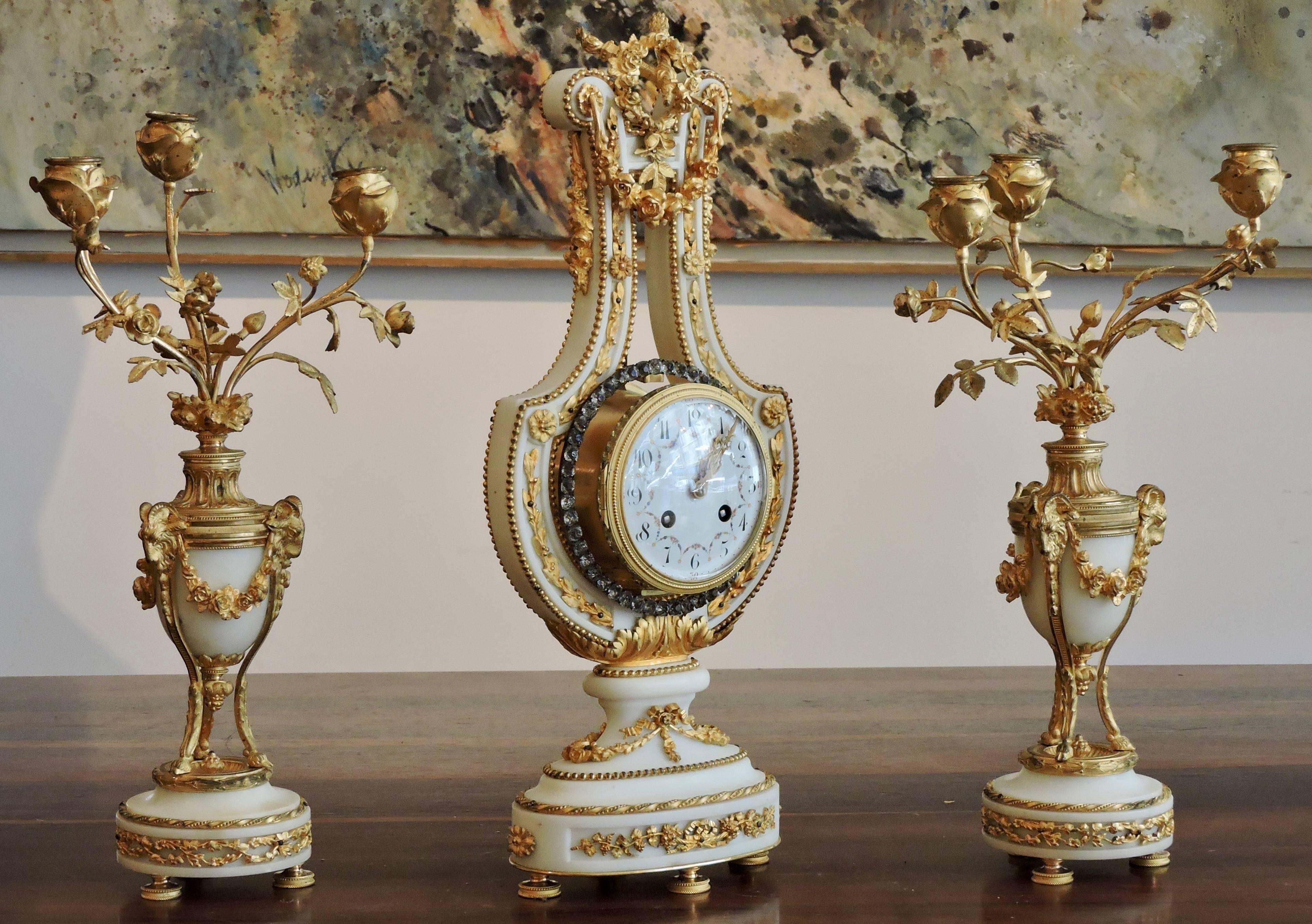 A 19th century French Ormolu and White Marble Three-Piece Lyre Clock Garniture
The central clock is shaped from white marble as a lyre, and mounted all over with gilt bronze leaves and swags, and jewellered torsades.
The pair of flanking candelabra