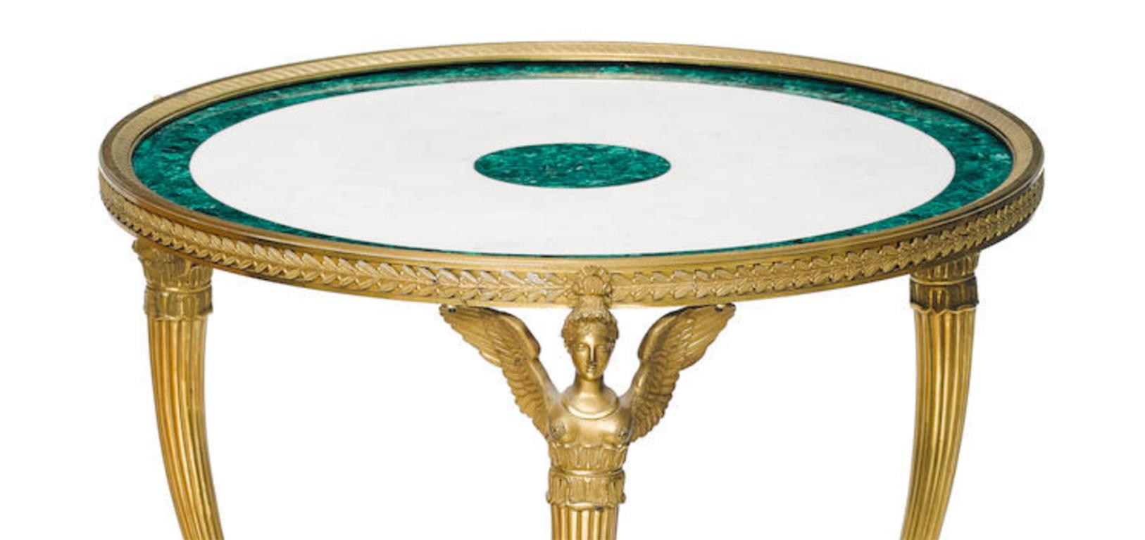 Table Guéridon en bronze doré, malachite et marbre blanc, de style Empire,
 vers 1870

Le plateau circulaire repose sur trois cariatides féminines ailées en bronze et se termine par des pieds en patte reliés par une civière tri-formée, sur des