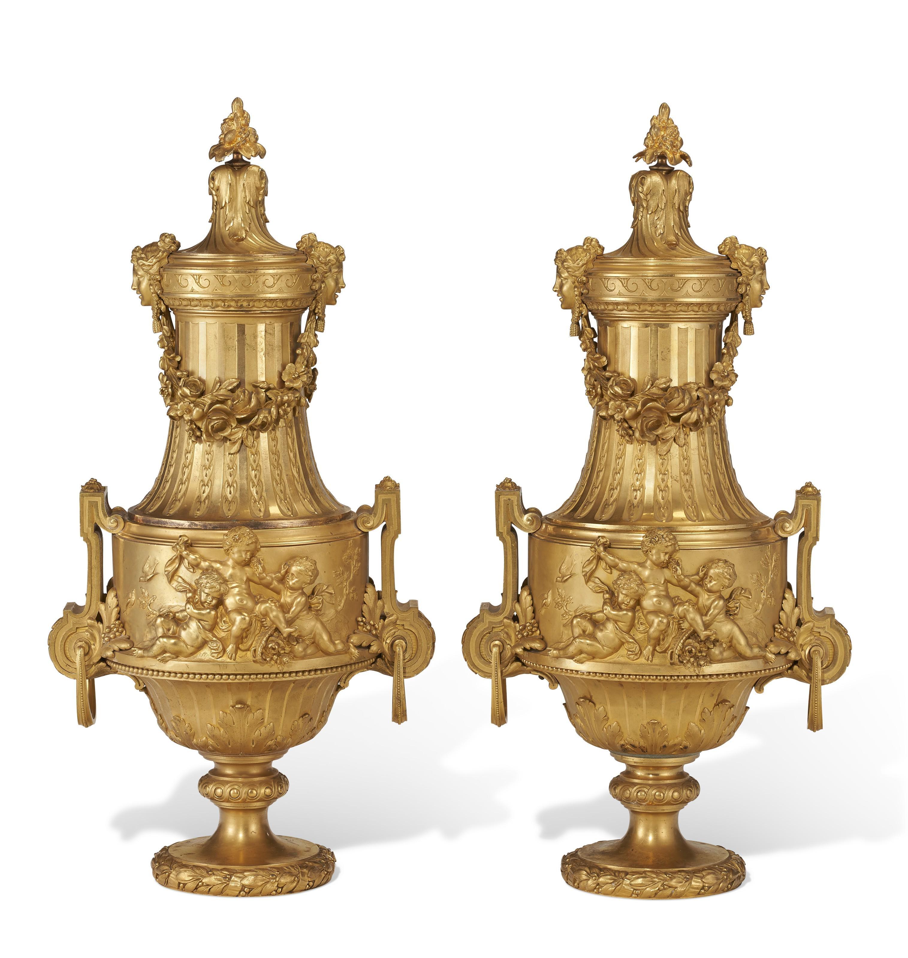 Our pair of exceptional ormolu bronze urns are attributed to French designer, Maxime Secretant, circa 1890s. They feature spiral-gadrooned necks flanked by female masks linked by suspended floral swags and bodies cast in relief with putti in