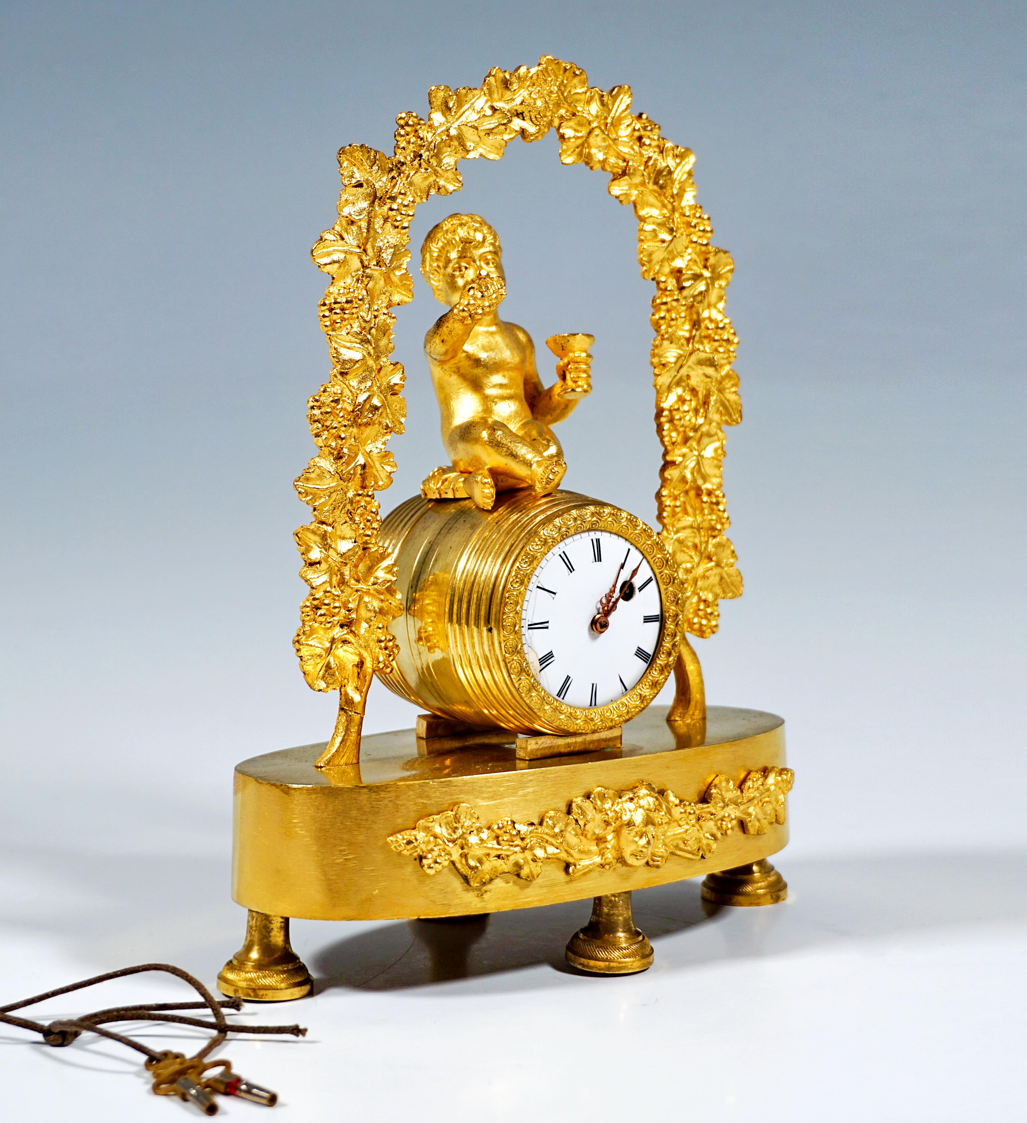 Fire-gilded bronze table clock: a childlike bacchanal seated on a wine barrel under an arched vine arbour, holding grapes and a wine glass in his hands, on an oval base with an appliquéd relief ornament on the front: crossed thyrsus rods entwined