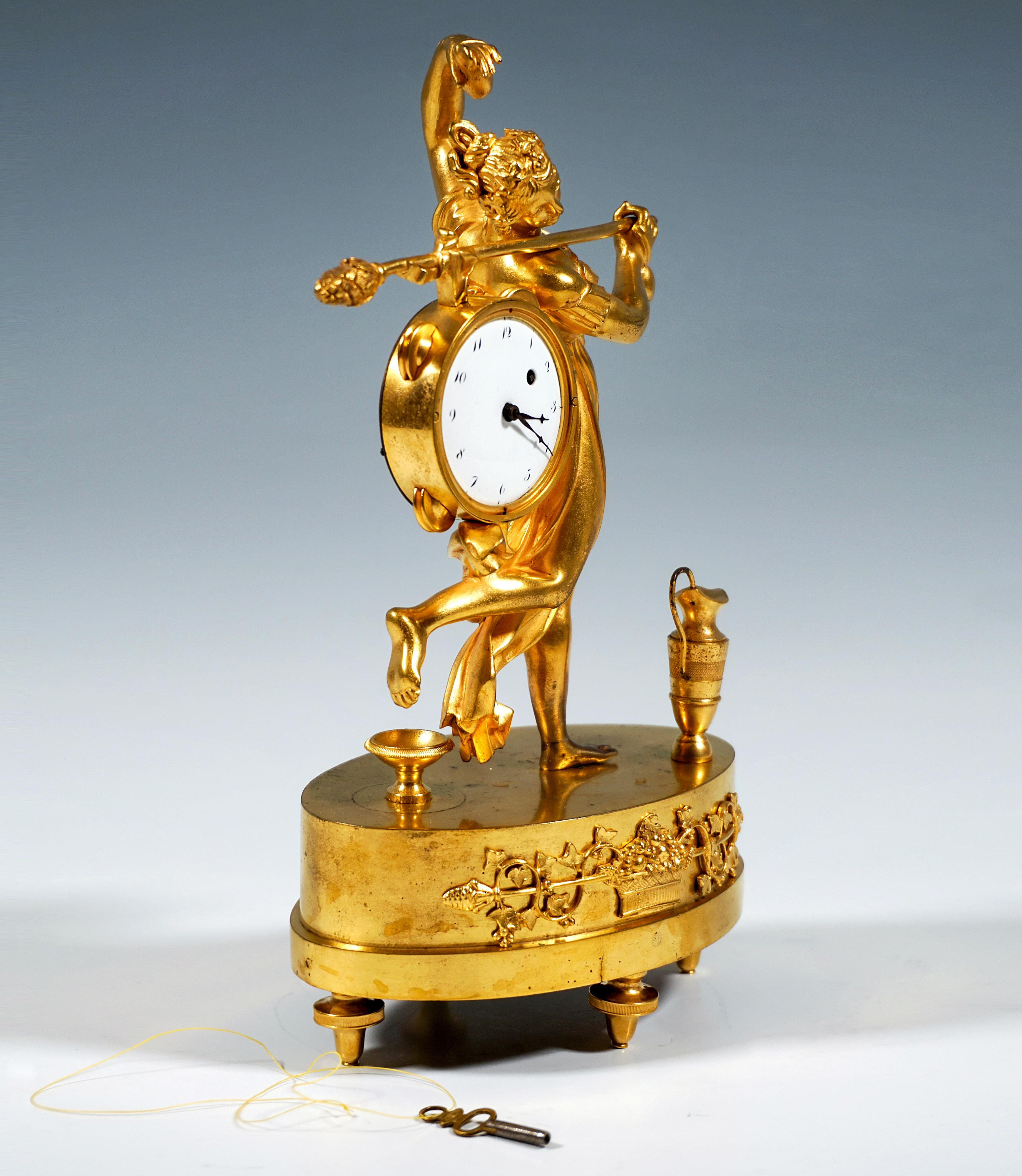 Fire-gilded bronze table clock: Dancing female figure on a high, tiered oval base with an appliquéd relief ornament on the front featuring a fruit basket and a Thyrsos staff entwined with vine leaves, on four fluted screw feet, the young woman,
