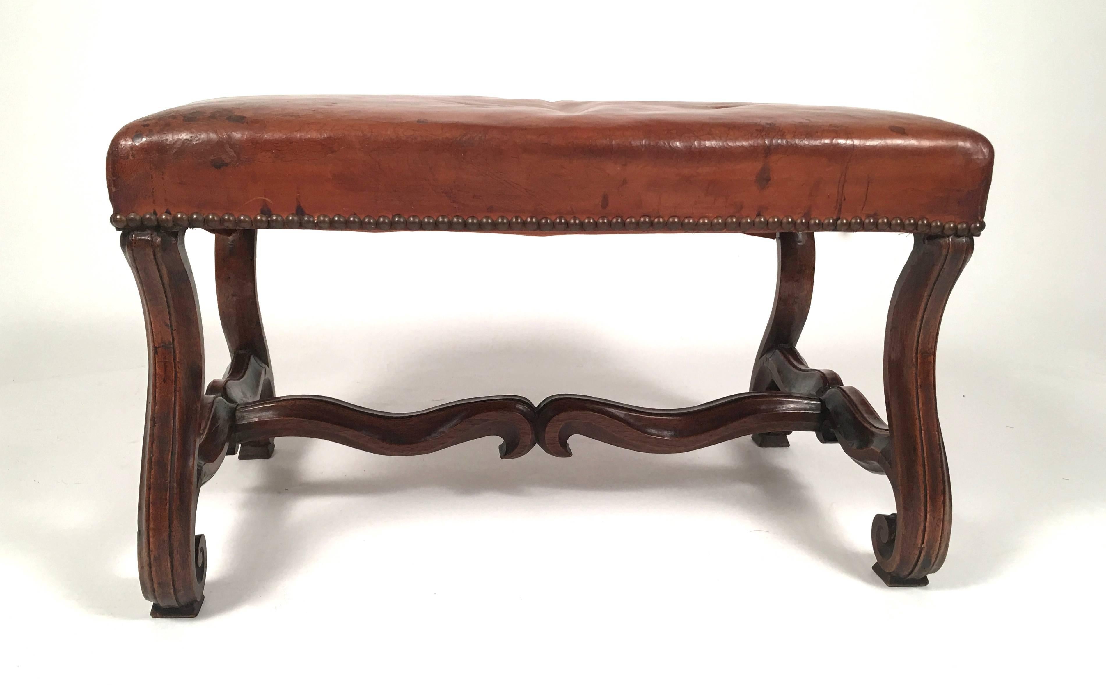 A French Louis XIV style Os de Mouton (literally sheep's bone) carved walnut ottoman, bench, or footstool with well patinated old leather upholstery and brass nail heads around the border, the S- curved legs joined by a serpentine shaped carved
