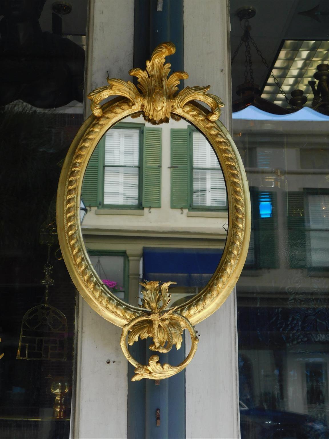 French oval gilt wood and gesso wall mirror with a foliage crest, gadrooned border, and single scrolled acanthus candle arm, Late 18th century. Mirror retains the original glass & wood backing.