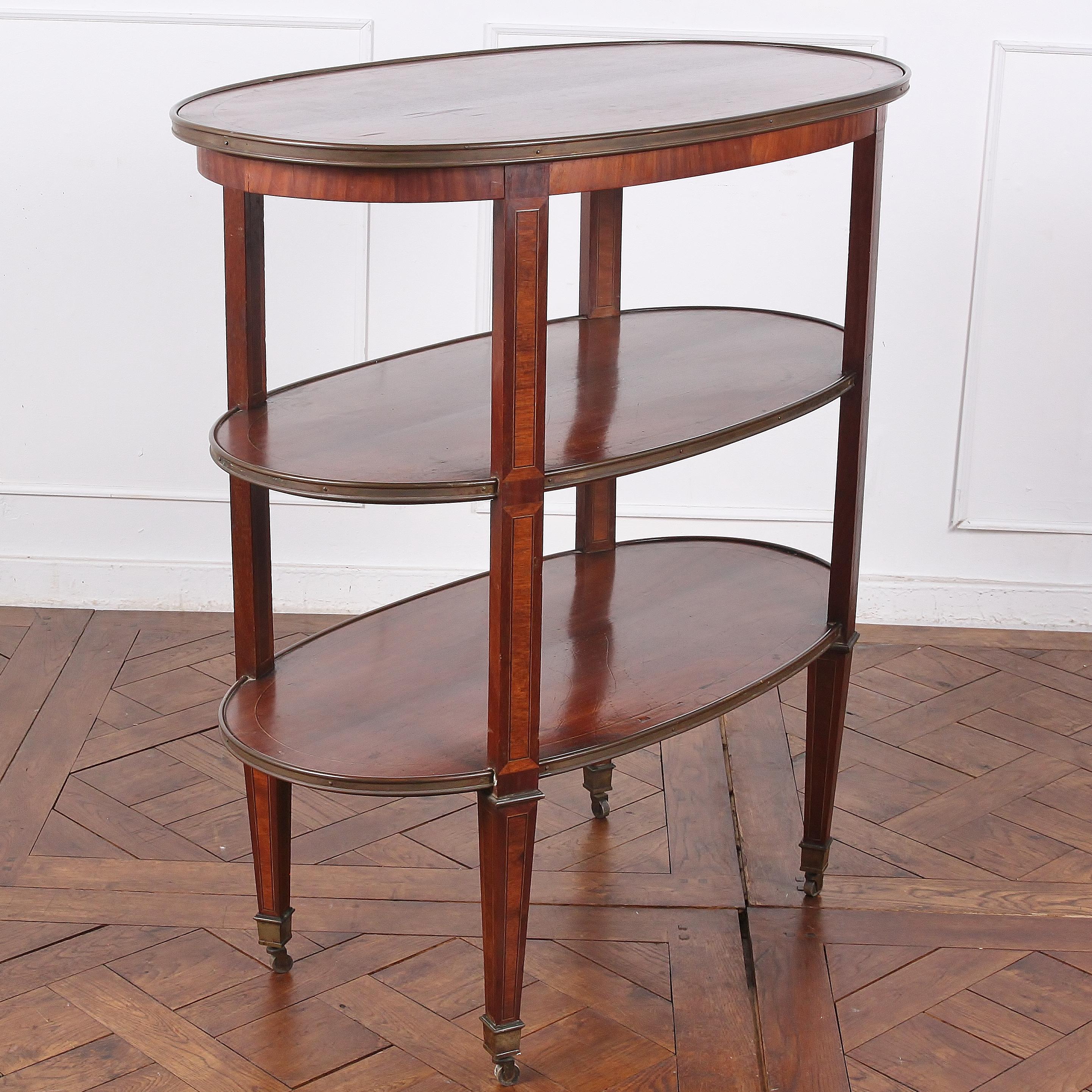 A three-tier oval mahogany trolley or stand with brass edge details, boxwood and walnut inlaid stringing, and retaining its original casters. Inlaid cross-banded panels to the faces of each leg. This is larger-scale than a typical trolley and could