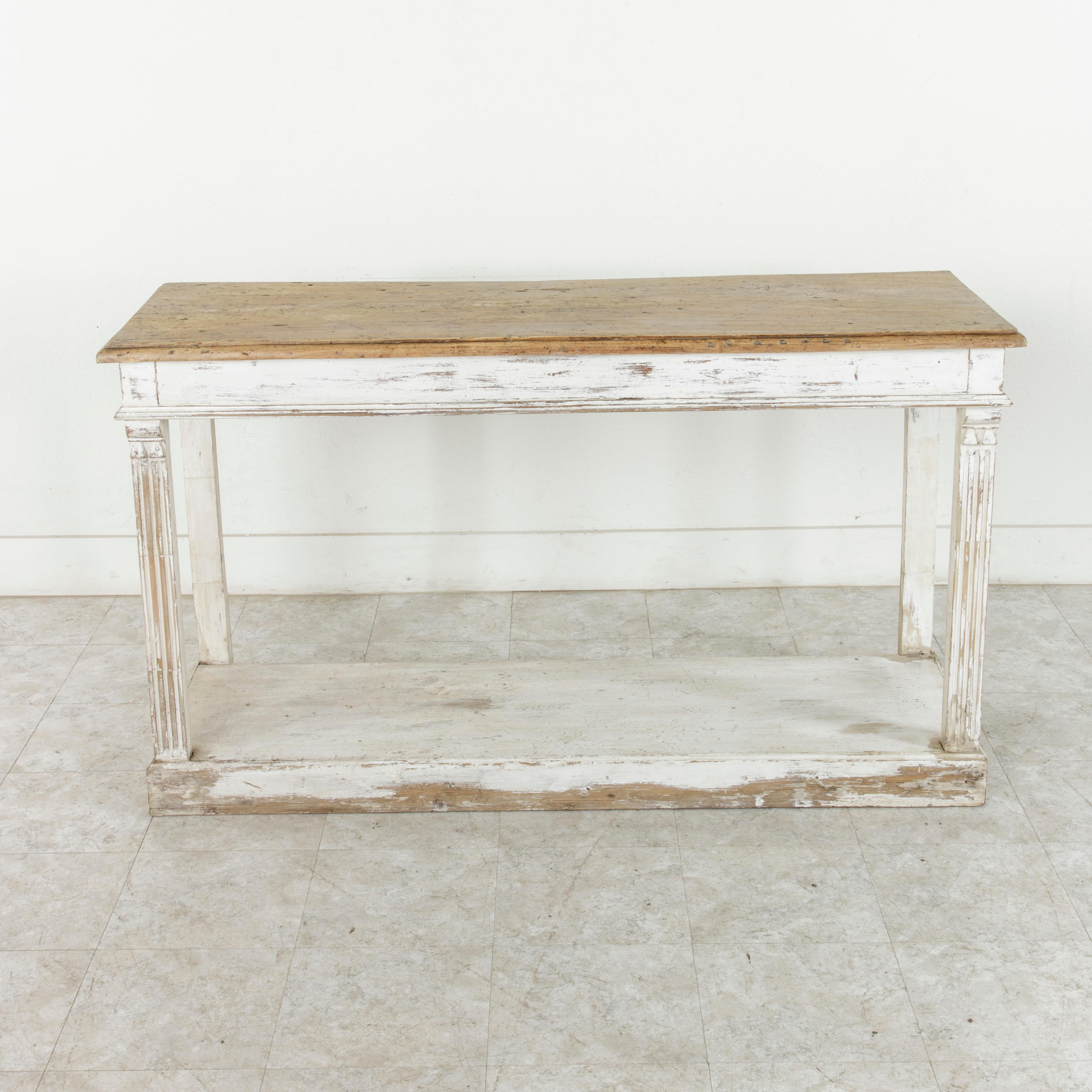 This French painted console table from the turn of the 20th century features a solid beveled walnut top made from a single plank of wood, naturally distressed from years of use. Supported by fluted columns at the front, the base is painted in white