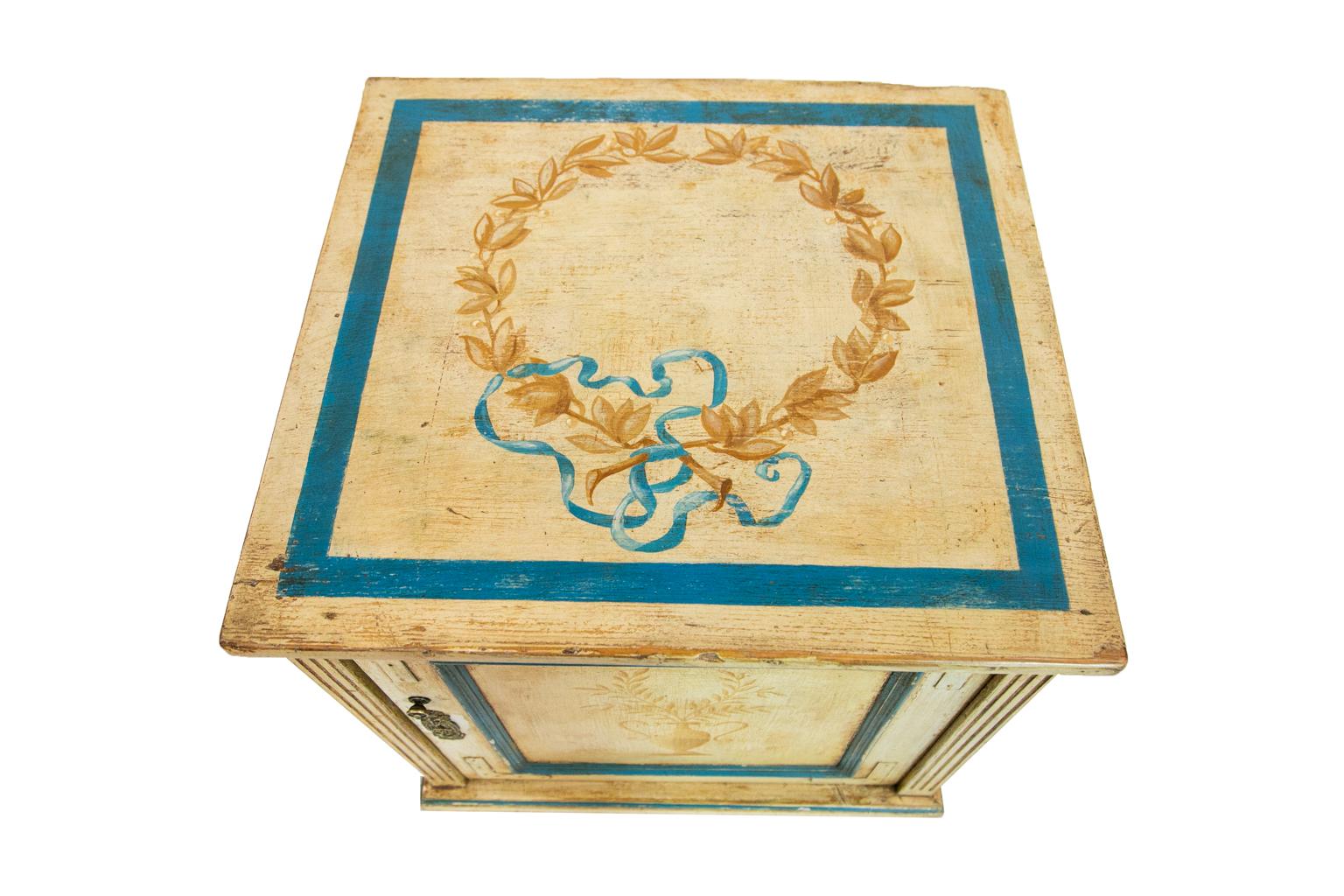 The top of this French cupboard is hand painted with a floral wreath tied with blue ribbon and framed with a blue painted square. The door is painted with a classical urn holding ferns. The door frame is reinforced with applied metal corners. The