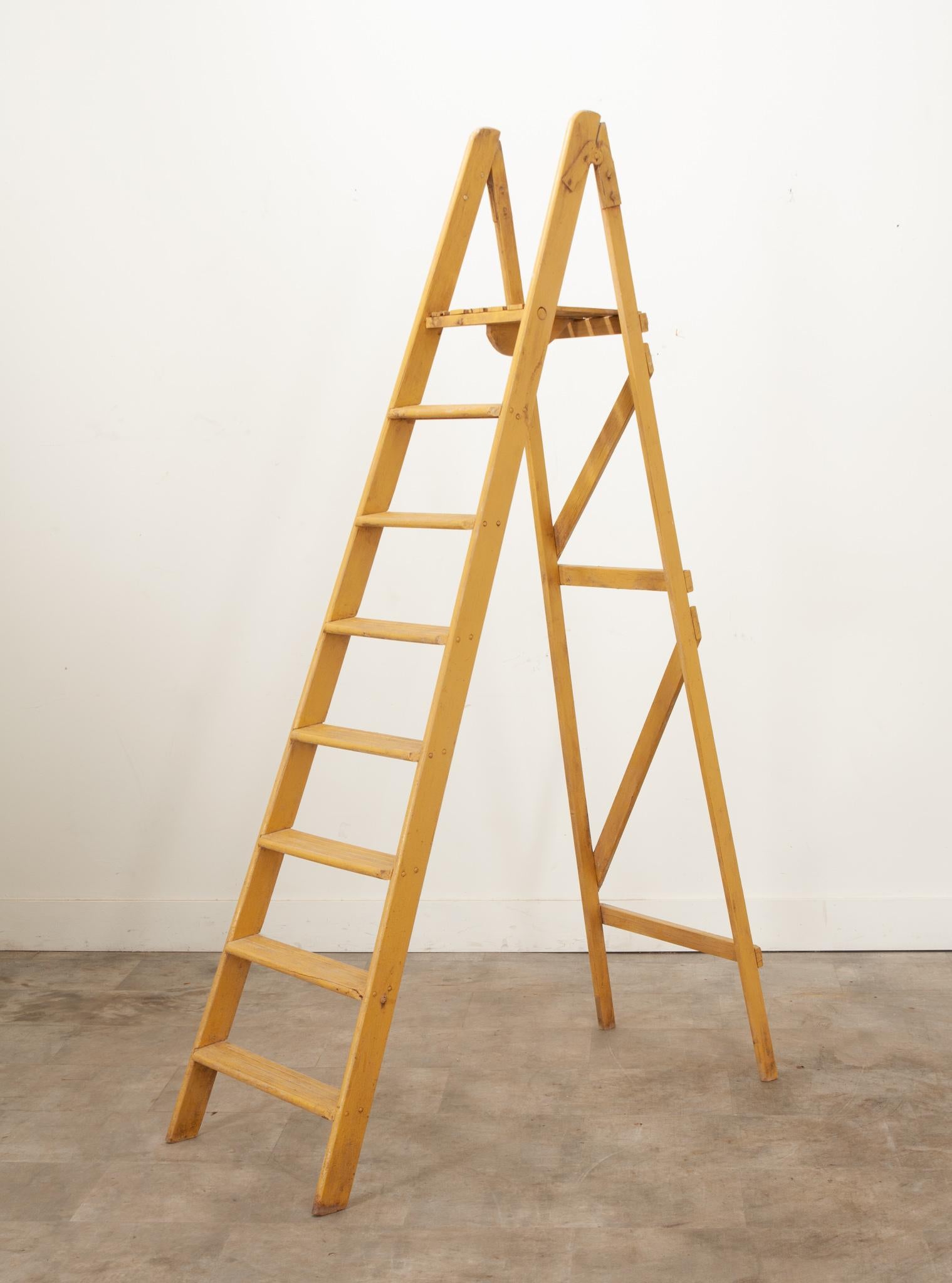 A French folding ladder, perfect for display or unique shelving. Painted yellow, this ladder is fitted with seven rungs, a slatted top platform for additional accessibility, and hinges that allow it to be folded and stored flat. This piece is not