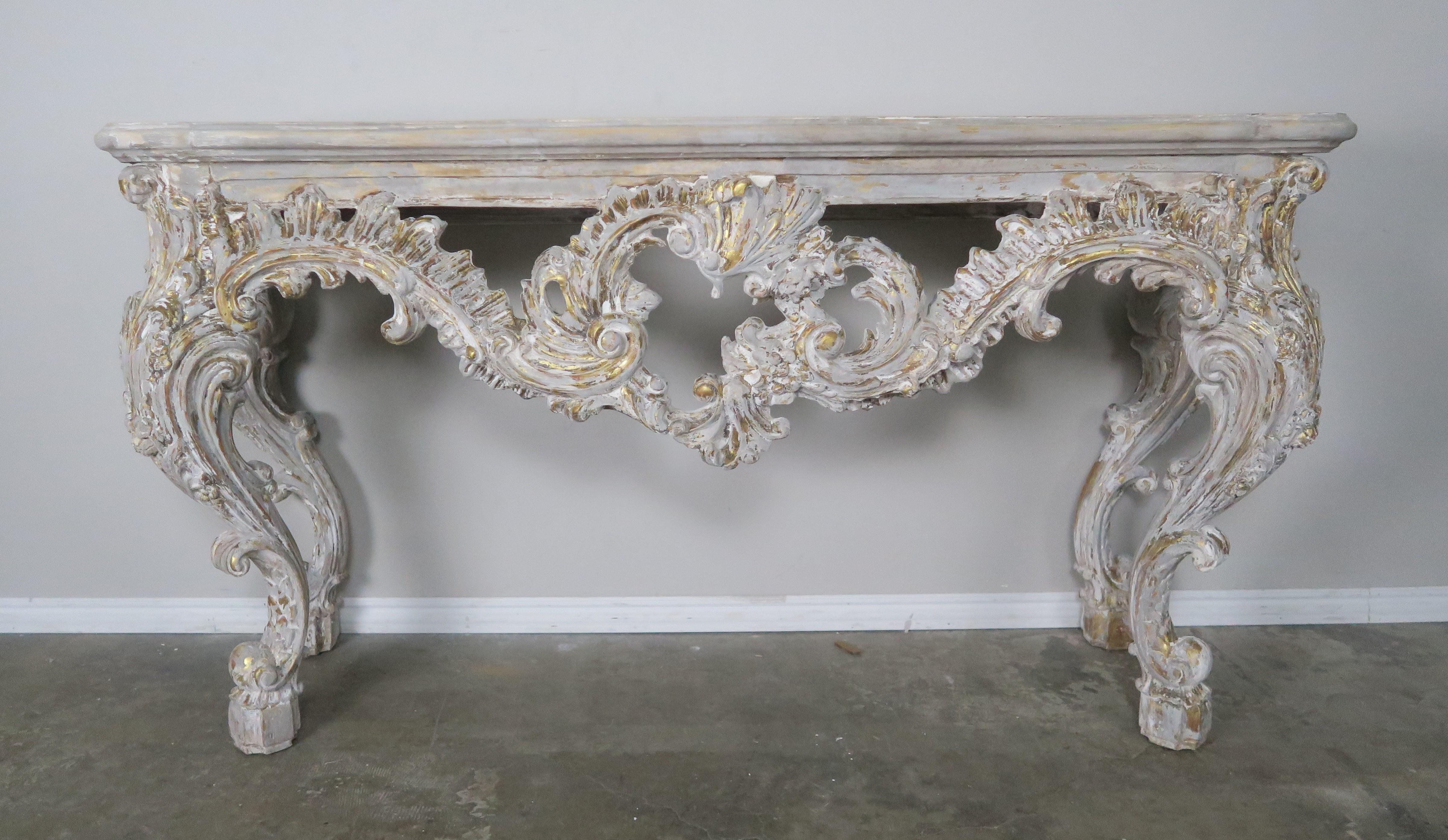 French heavily carved Louis XV style console painted in an antique white coloration with gold gilt highlights throughout. The console stands on four cabriole legs that end in ram's head feet. The console is beautifully carved with swirling acanthus