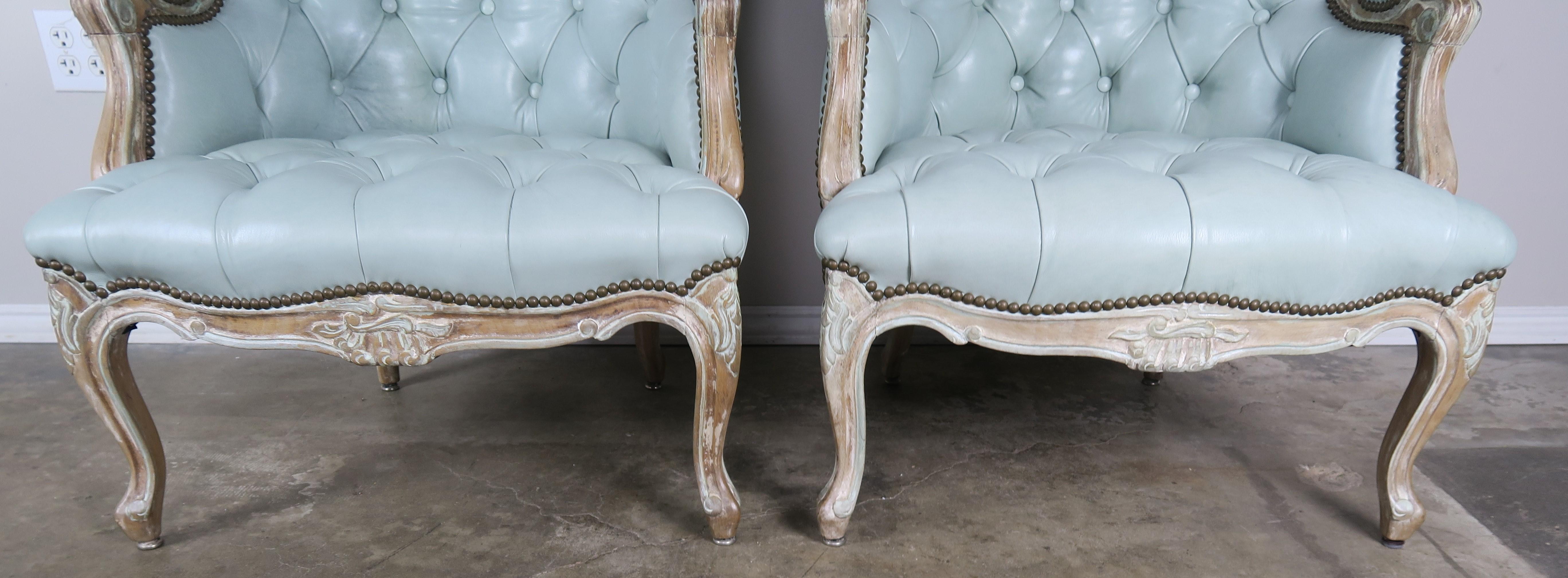 Pair of 19th century French painted Louis XV style bergere armchairs upholstered in a soft blue colored tufted leather with brass nailhead trim detail. The armchairs stand on four cabriole legs with rams head feet. Beautiful distressed painted