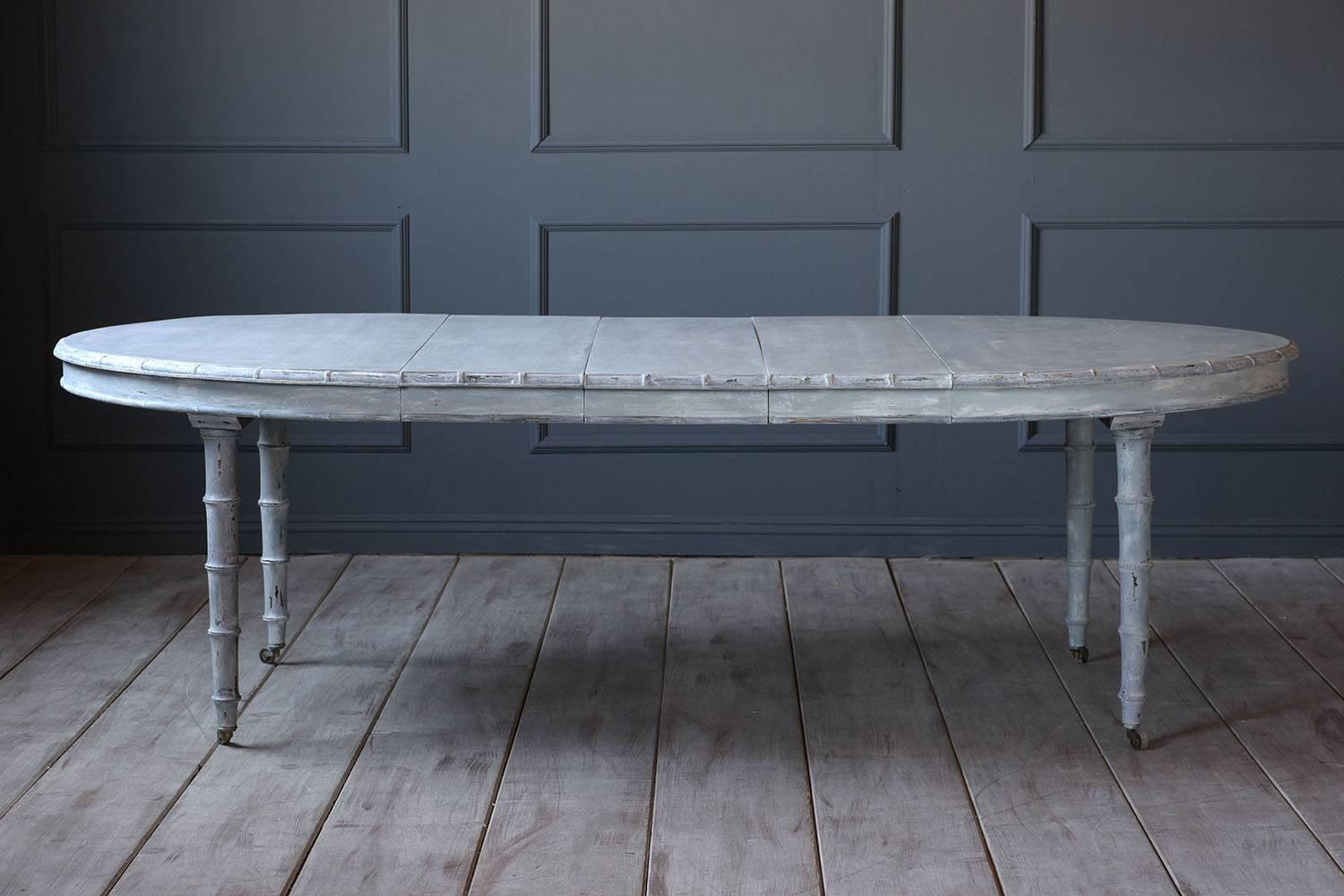 This 1900s French chinoiserie style oval walnut wood dining table is painted in a pale gray and off-white with a distressed finish. The edges and legs of the table have faux bamboo carved accents. The table is accompanied by the original three