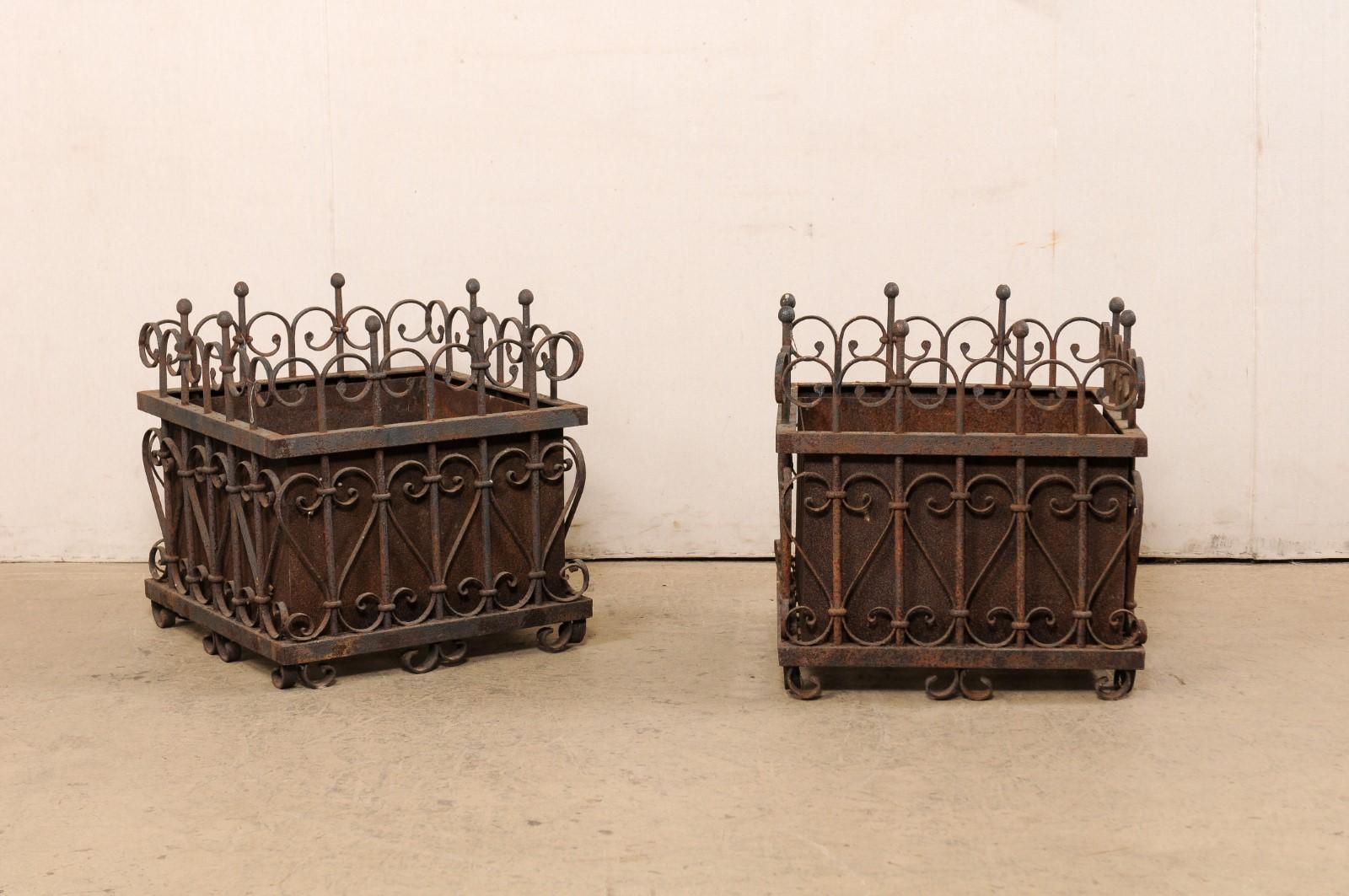 A French pair of wrought iron garden planters from the early 20th century. This antique pair of planters from France, each approximately two feet square in shape, feature scrolled ornamental wrought iron work, with petite ball-shaped finials,