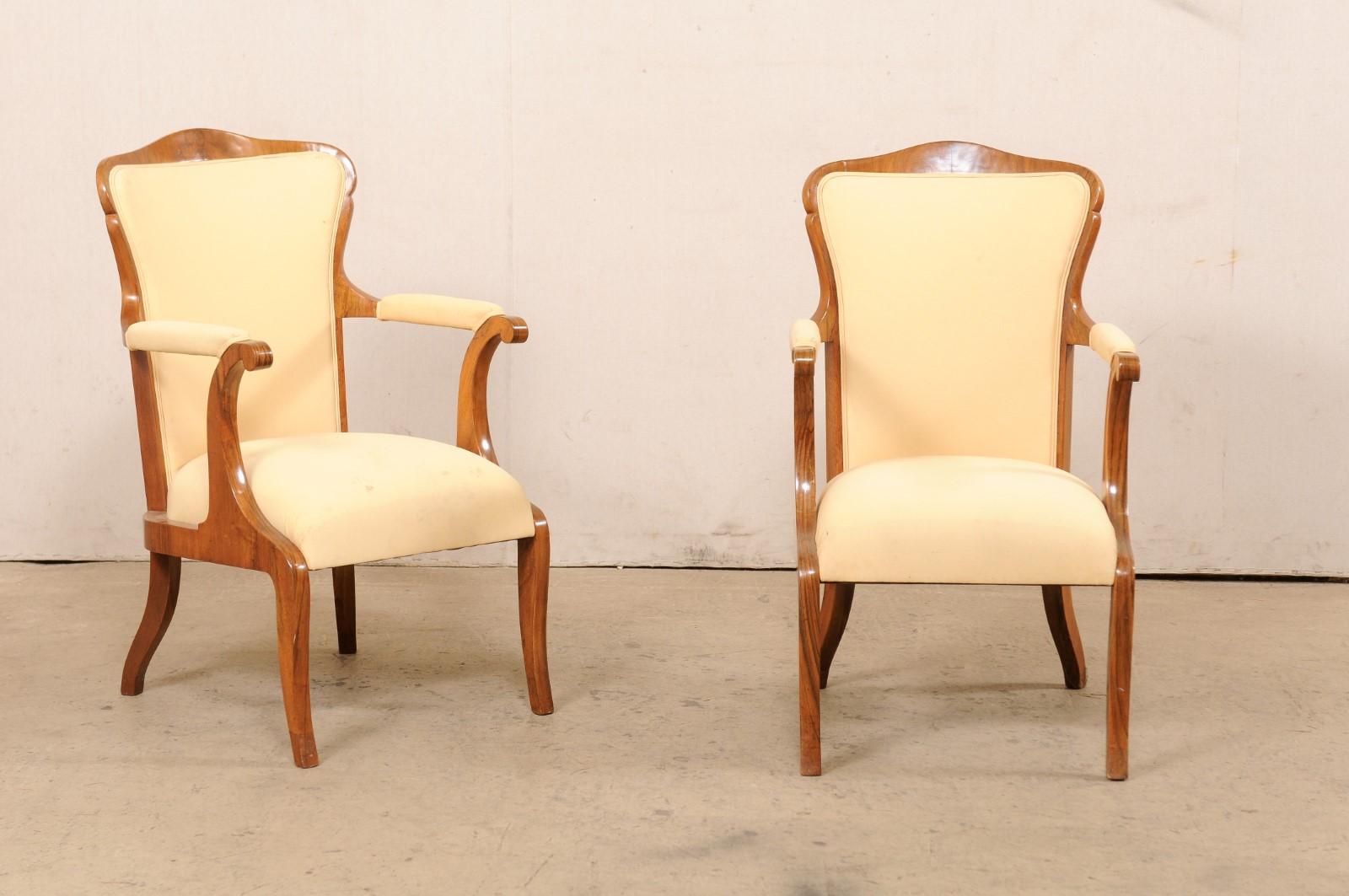 A French pair of carved-wood and upholstered fauteuil armchairs from the early to mid 20th century. This pair of armchairs from France have upholstered backs framed an elegantly carved and arched wooden top-rail, with softly rounded shoulders, that