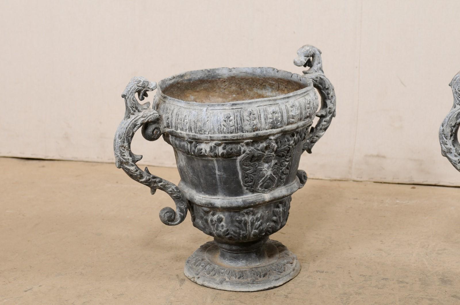 A French pair of decorative urn planters from the 18th century. This antique pair of lead garden ornaments from France stand approximately 16 inches in height, each featuring an urn raised on a rounded pedestal, leaf motif decor, and s-scroll