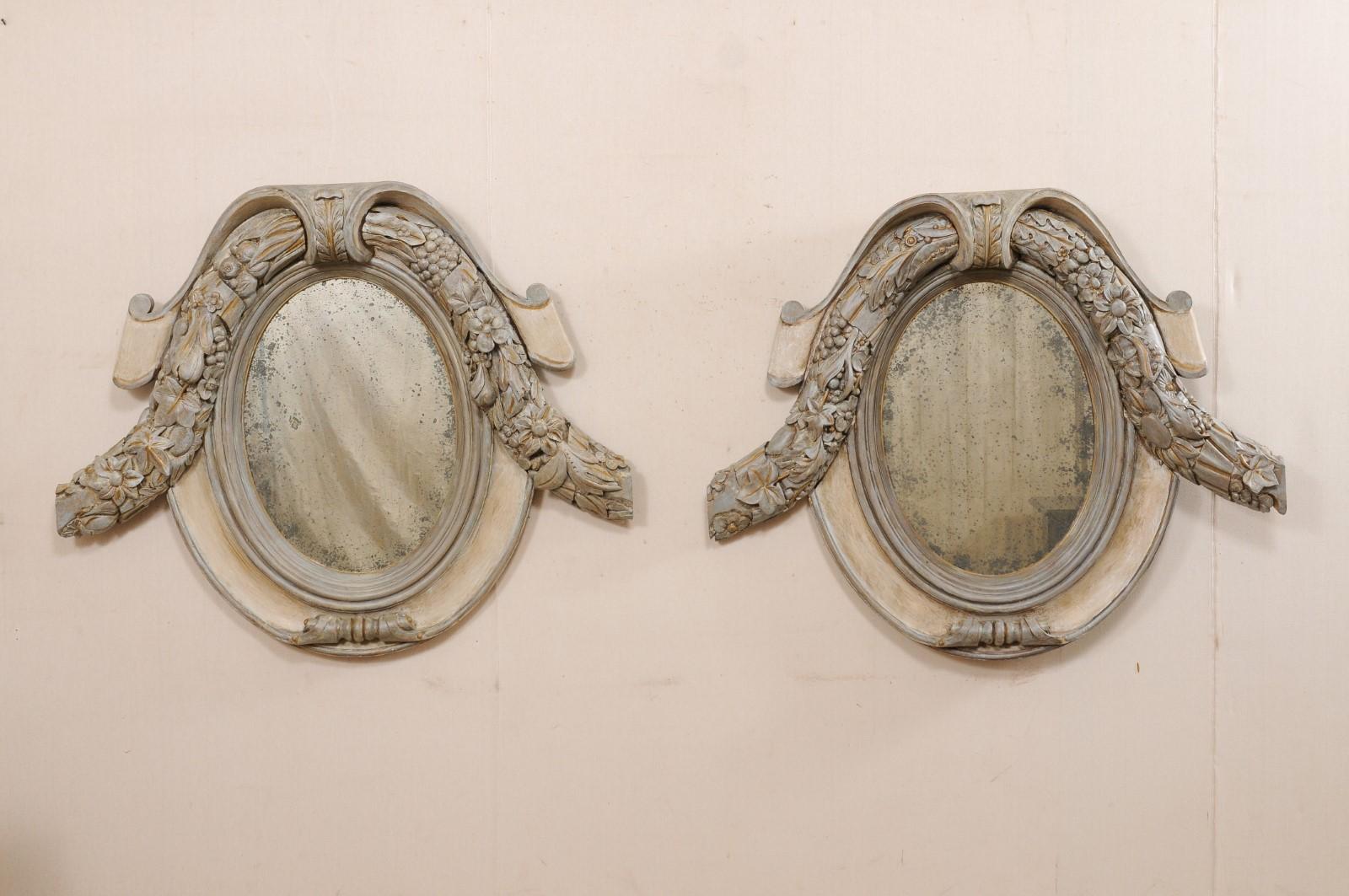 A French pair of cartouche and floral carved mirrors from the early 20th century. This antique pair of mirrors from France have beautifully carved cartouche inspired frames, mostly oval in shape, with a leaf and floral carved swag garland wrapped